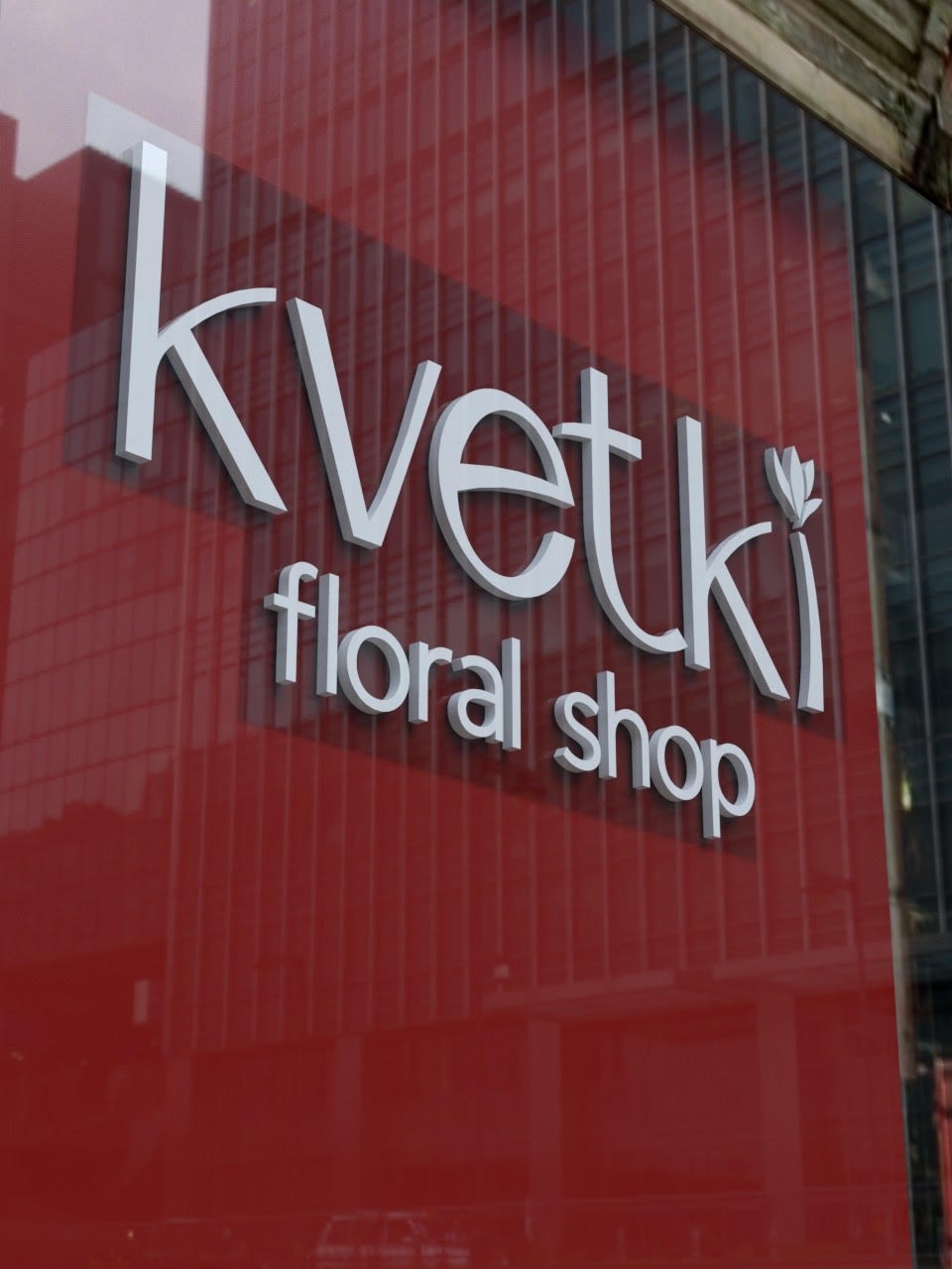 Elegancki szyld 3D w kolorze białym na czerwonym tle, z napisem 'Kvetki floral shop', umieszczony na szklanej fasadzie budynku, odbijający miejski krajobraz.