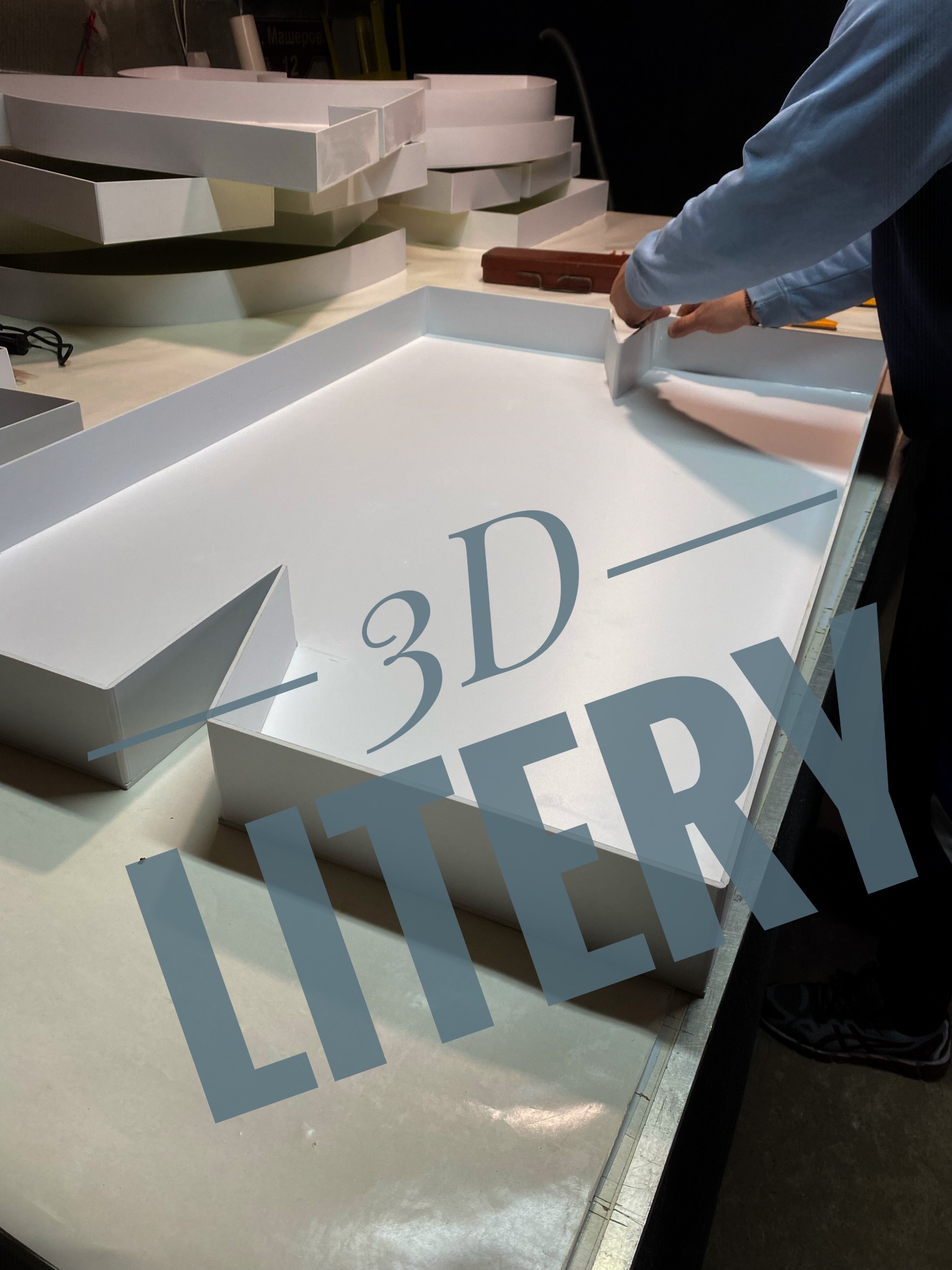 Ręczne składanie białych liter 3D na stole warsztatowym, widoczne fragmenty innych liter i narzędzia. Oświetlenie studyjne, praca w toku.