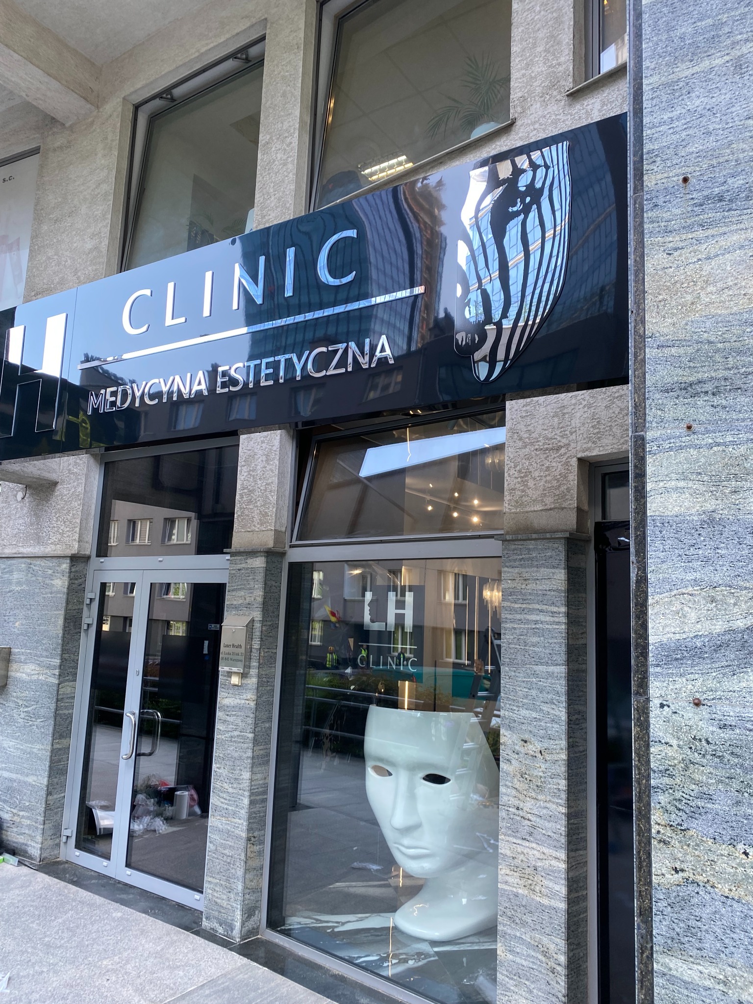 Elegancki szyld kliniki medycyny estetycznej z chromowanymi literami i graficznym logo, umieszczony nad wejściem do budynku z marmurową fasadą i witryną.