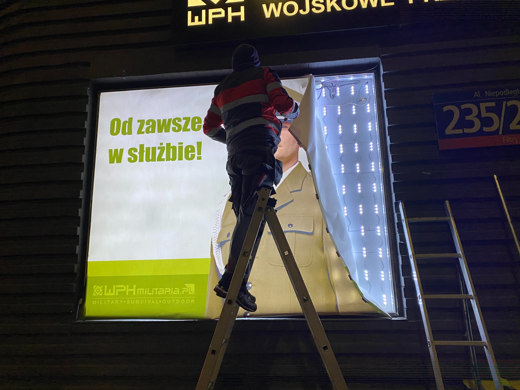 Montaż podświetlanego baneru reklamowego z hasłem 'Od zawsze w służbie!' i logotypem WPH MILITARIA.PL, widoczne drabiny i pracownik podczas instalacji.