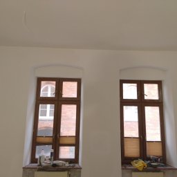 BEC-BUD FIRMA USŁUGOWO-HANDLOWA KAROL ZAGDAN - Białe ściany po malowaniu z dwoma drewnianymi oknami. Na parapecie widoczne narzędzia malarskie i akcesoria. Minimalistyczne wnętrze po remoncie.