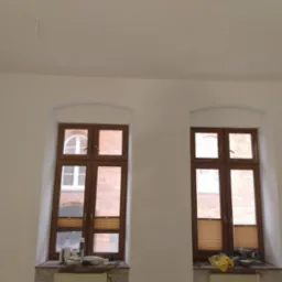 Białe ściany po malowaniu z dwoma drewnianymi oknami. Na parapecie widoczne narzędzia malarskie i akcesoria. Minimalistyczne wnętrze po remoncie.