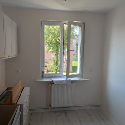 BEC-BUD FIRMA USŁUGOWO-HANDLOWA KAROL ZAGDAN - Jasna kuchnia w trakcie montażu: białe szafki z złotymi uchwytami, otwarte okno z widokiem na zieleń, grzejnik pod oknem, panele podłogowe.