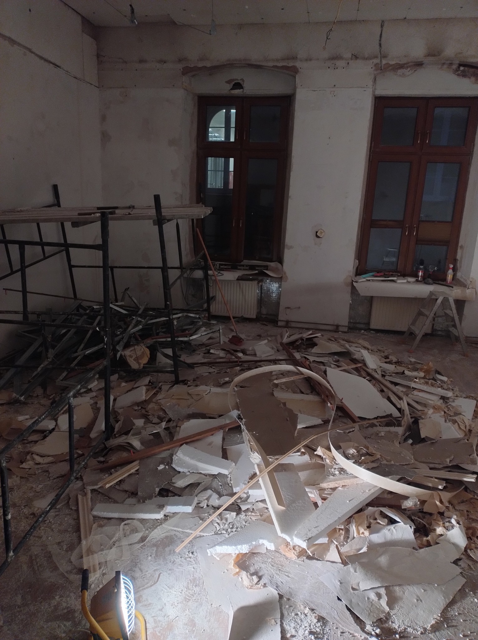 Demontaż płyty karton gips w pokoju z oknami. Widoczne rusztowanie, lampa budowlana i gruz. W tle grzejniki i drabina.