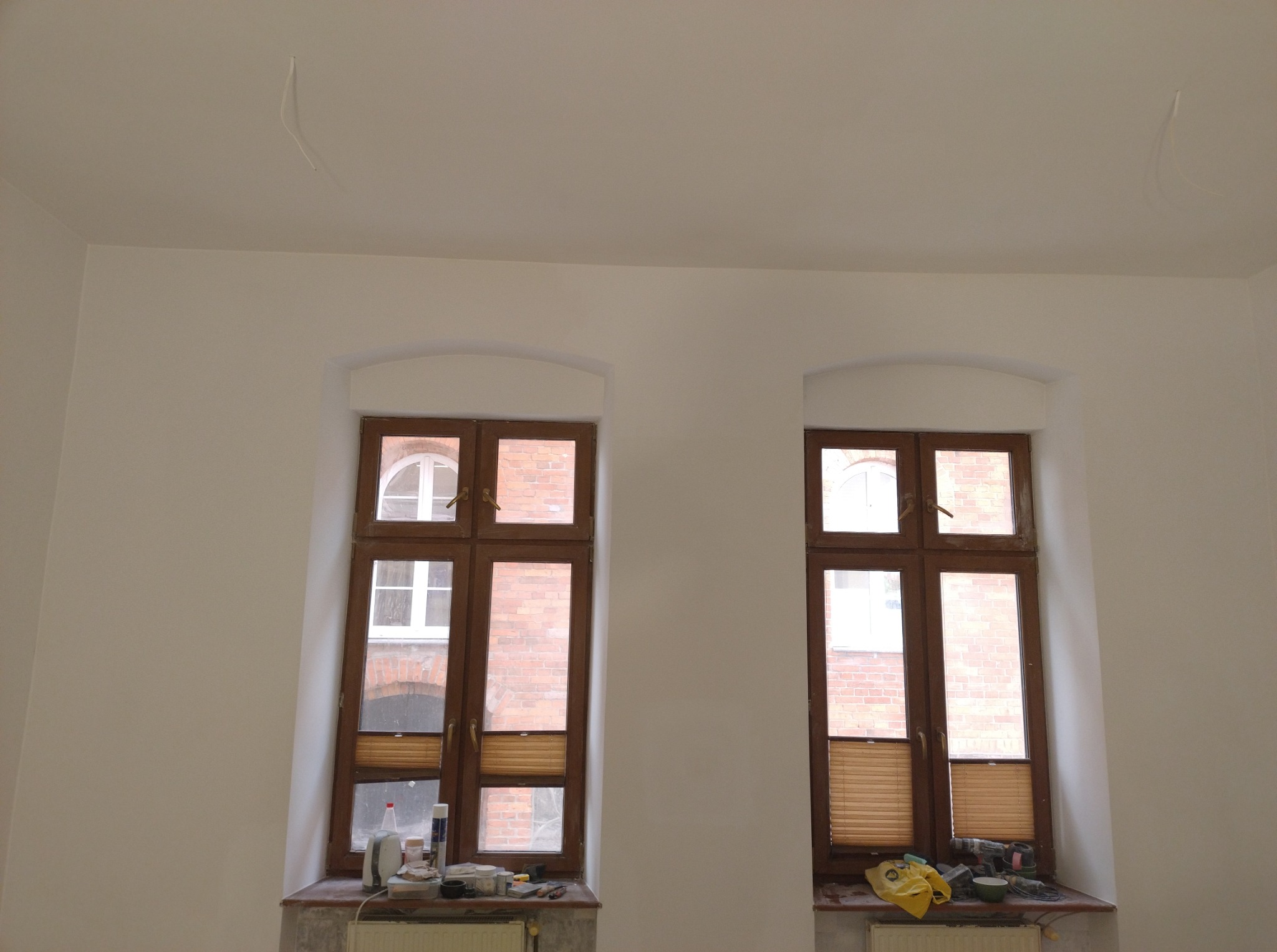 Białe ściany po malowaniu z dwoma drewnianymi oknami. Na parapecie widoczne narzędzia malarskie i akcesoria. Minimalistyczne wnętrze po remoncie.