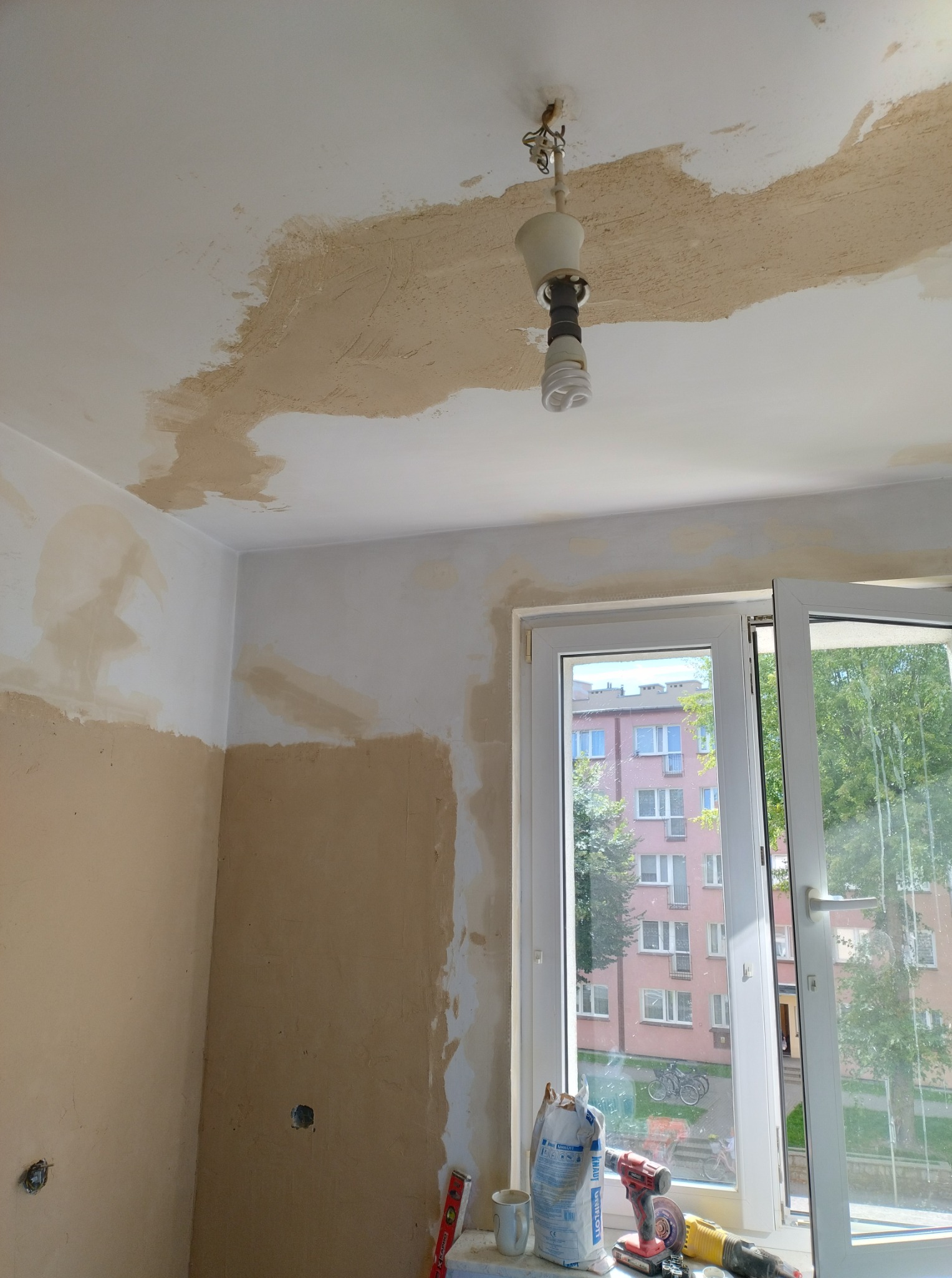 Wnętrze w trakcie szpachlowania: ściany częściowo pokryte gładzią, widoczne narzędzia i materiały budowlane na parapecie okna. W tle blok mieszkalny.