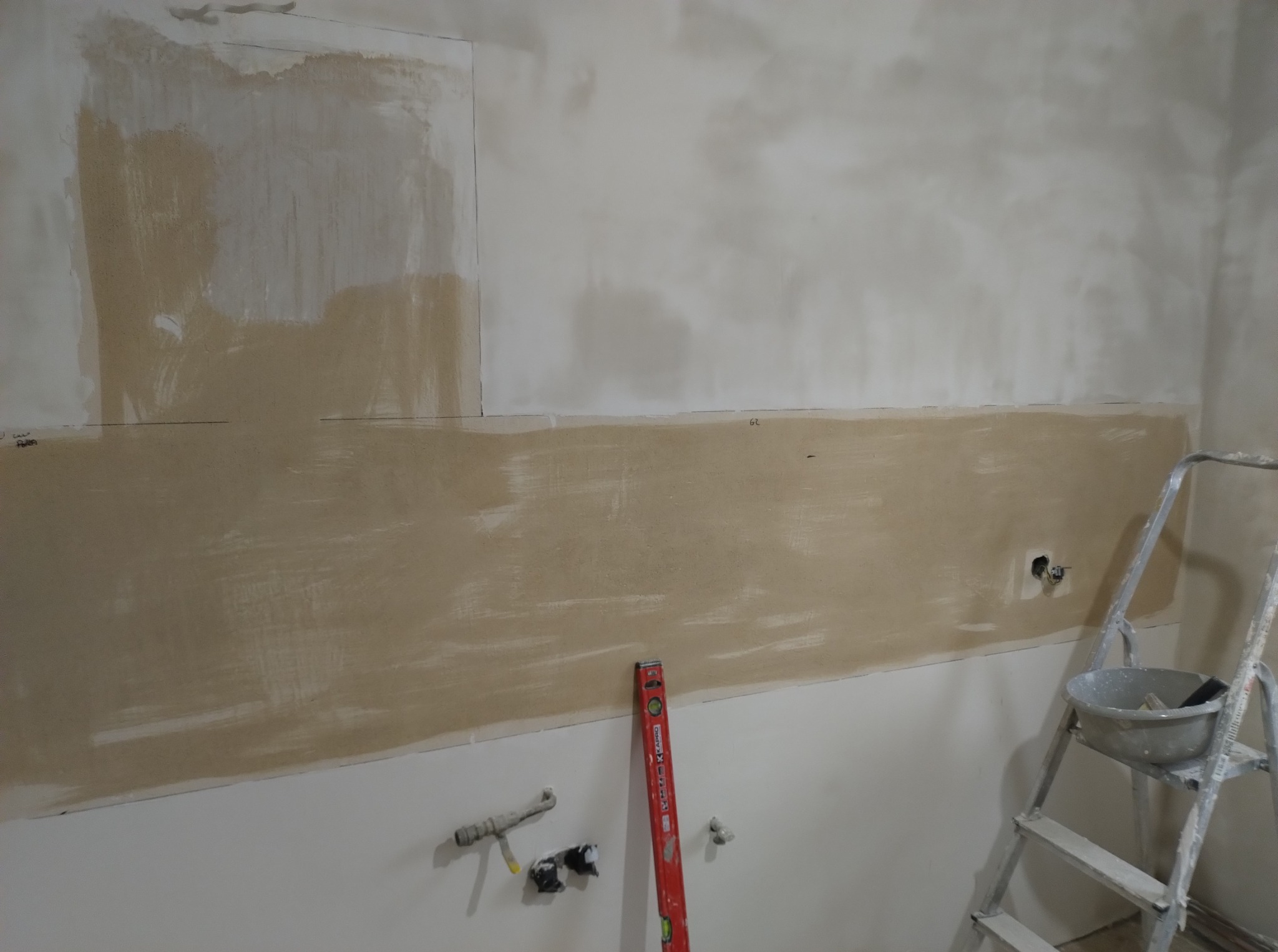 Ściana w trakcie gruntowania pod płytki w kuchni. Widoczny poziomica, drabina i puszka elektryczna. Częściowo zaszpachlowana ściana.