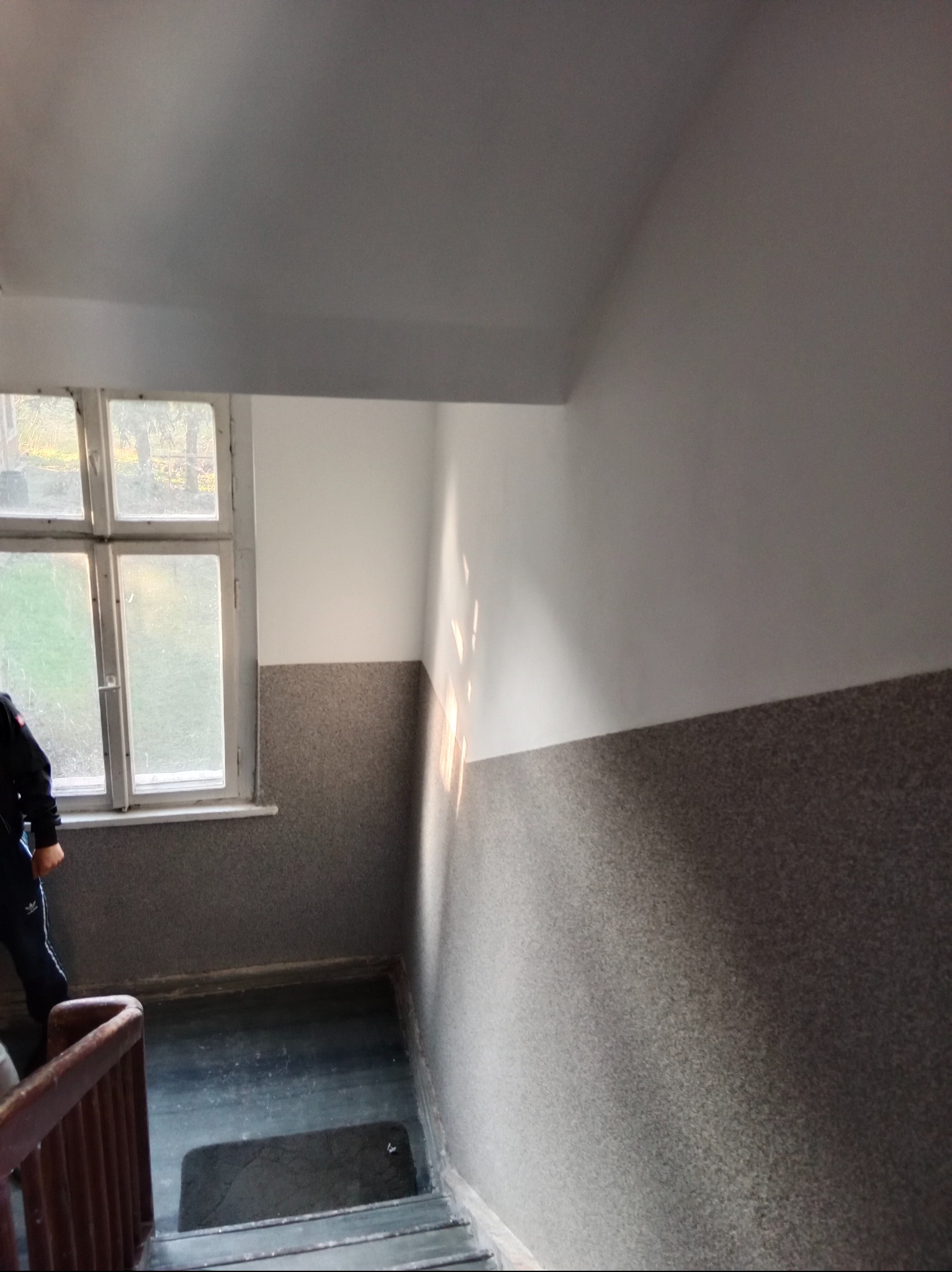 Klatka schodowa po remoncie: szare ściany do połowy wysokości, białe powyżej. Okno z widokiem na zieleń. Osoba w sportowym stroju schodzi po schodach.