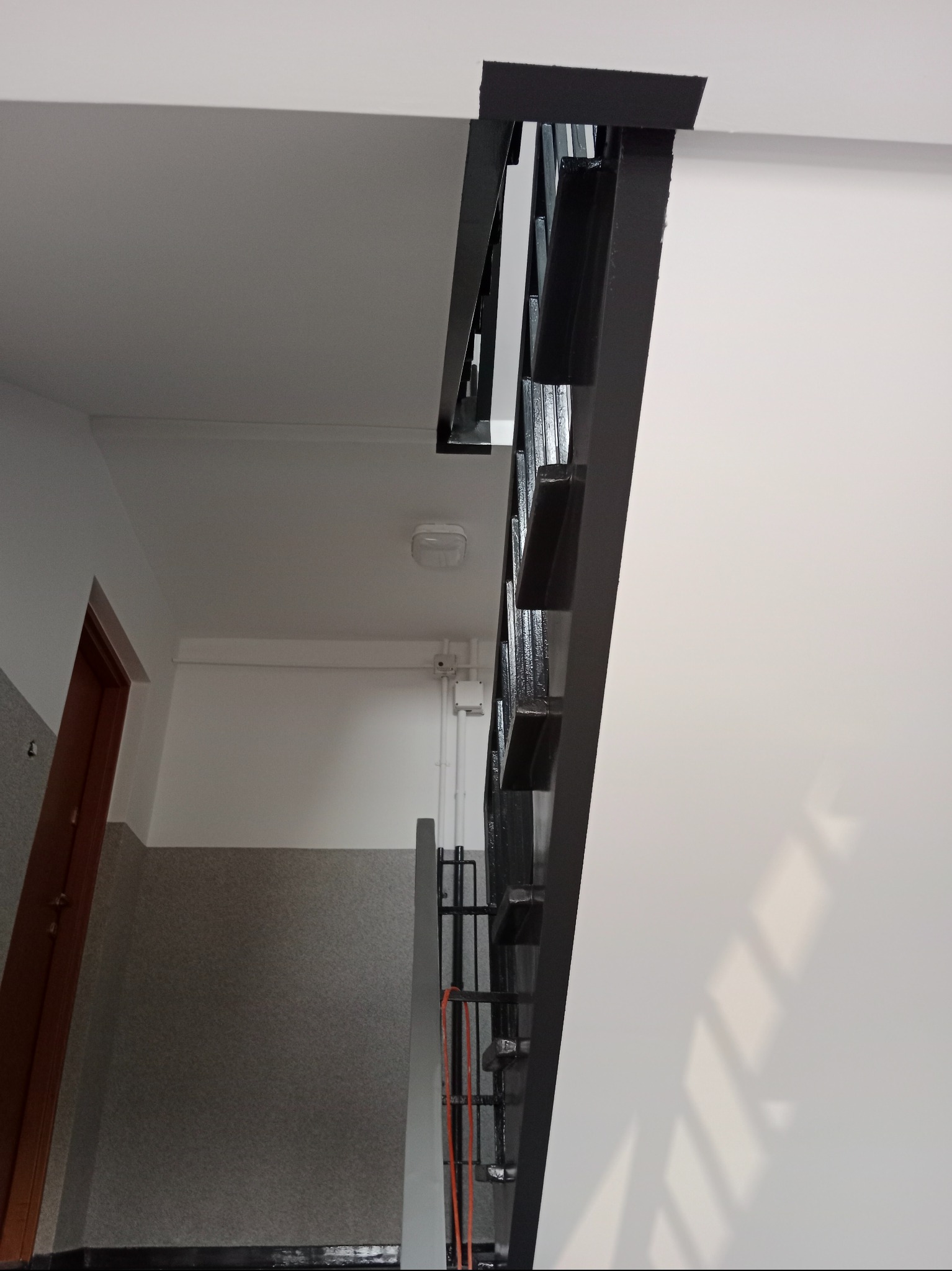 Klatka schodowa po remoncie: białe ściany, szare lamperie i czarna, metalowa balustrada. Widoczny fragment drzwi i sufit z oświetleniem. Ujęcie z dołu.