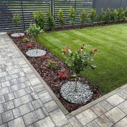 Michał Kowaliński - Zadbany ogród z równo przystrzyżonym trawnikiem, ozdobnymi drzewkami z czerwonymi akcentami, wysypanymi kamieniami i korą, oraz nowoczesnym, szarym ogrodzeniem.