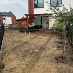 Przygotowany teren pod trawnik w Koleczkowie. Siatka wzmacniająca, młode drzewka wzdłuż ogrodzenia. W tle dom z pergolą i elementy wyposażenia ogrodu.