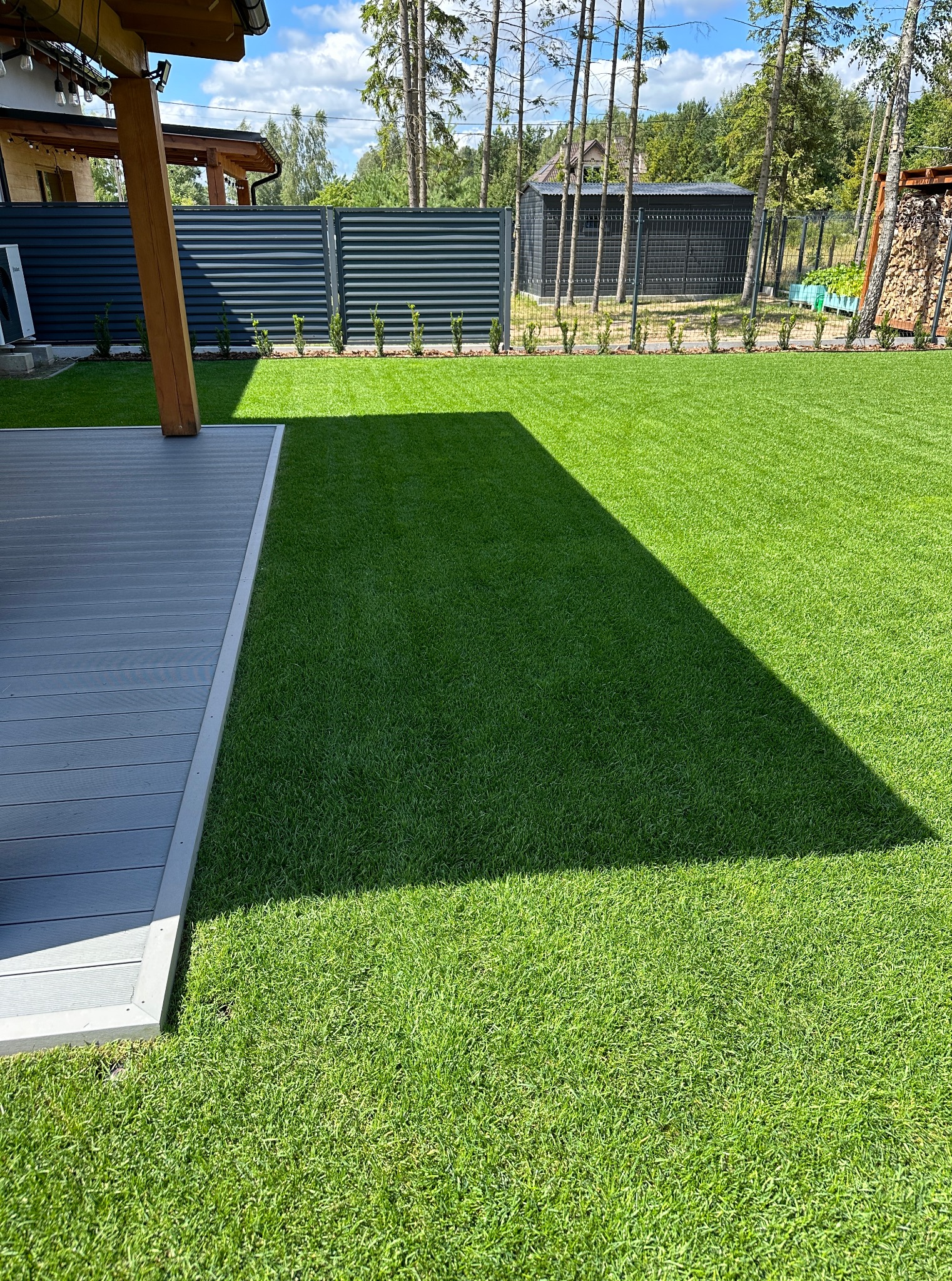 Idealnie przystrzyżony trawnik z równą linią cienia, graniczący z szarą deską tarasową i metalowym ogrodzeniem, otoczony zielenią. Perfekcyjna pielęgnacja ogrodu.
