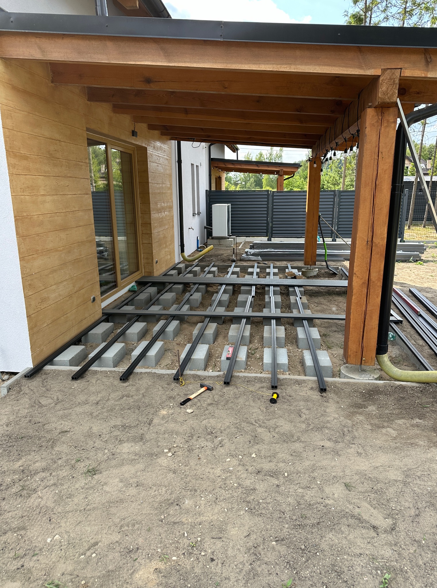 Montaż konstrukcji tarasu z belek stalowych na betonowych bloczkach, widoczne narzędzia, drewniana pergola w tle, dom z drewnianą elewacją.