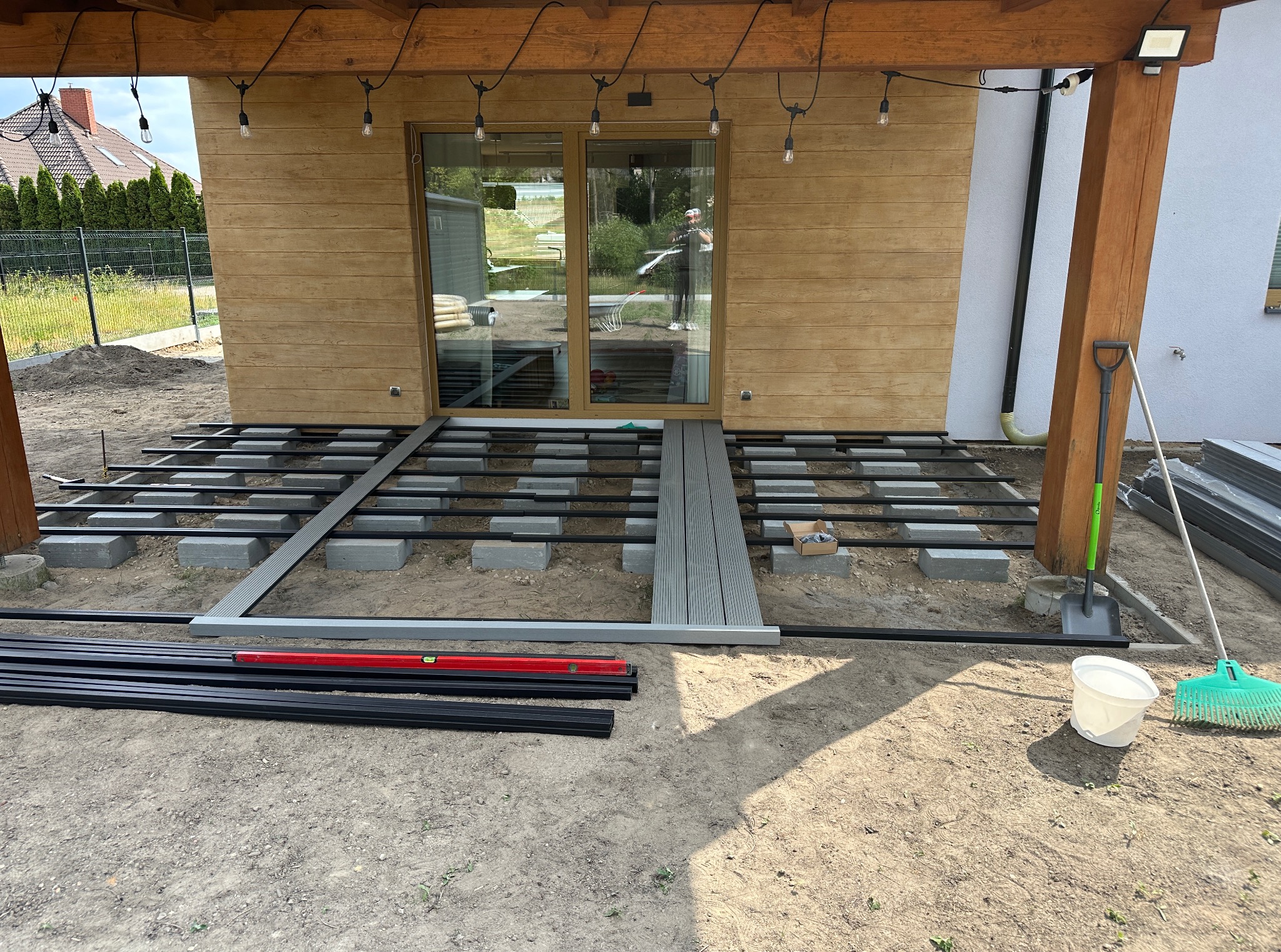 Montaż tarasu kompozytowego: widok na przygotowany stelaż z belek i bloczków betonowych, z częściowo ułożonymi deskami. Widoczne narzędzia i materiały budowlane.