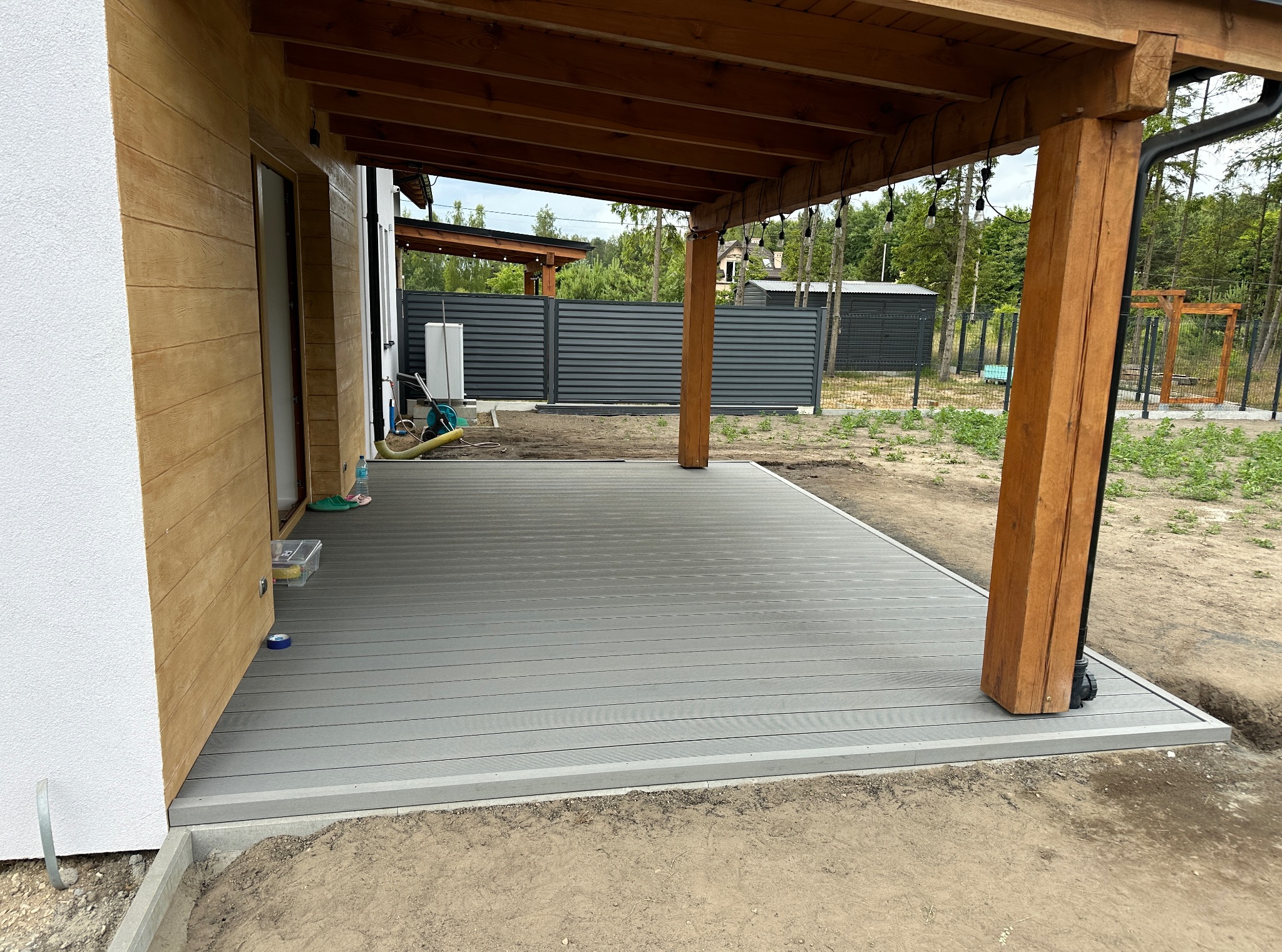 Nowoczesny taras z kompozytowych desek w odcieniach szarości, z drewnianą konstrukcją zadaszenia i widokiem na zadbany ogród z szarym ogrodzeniem.