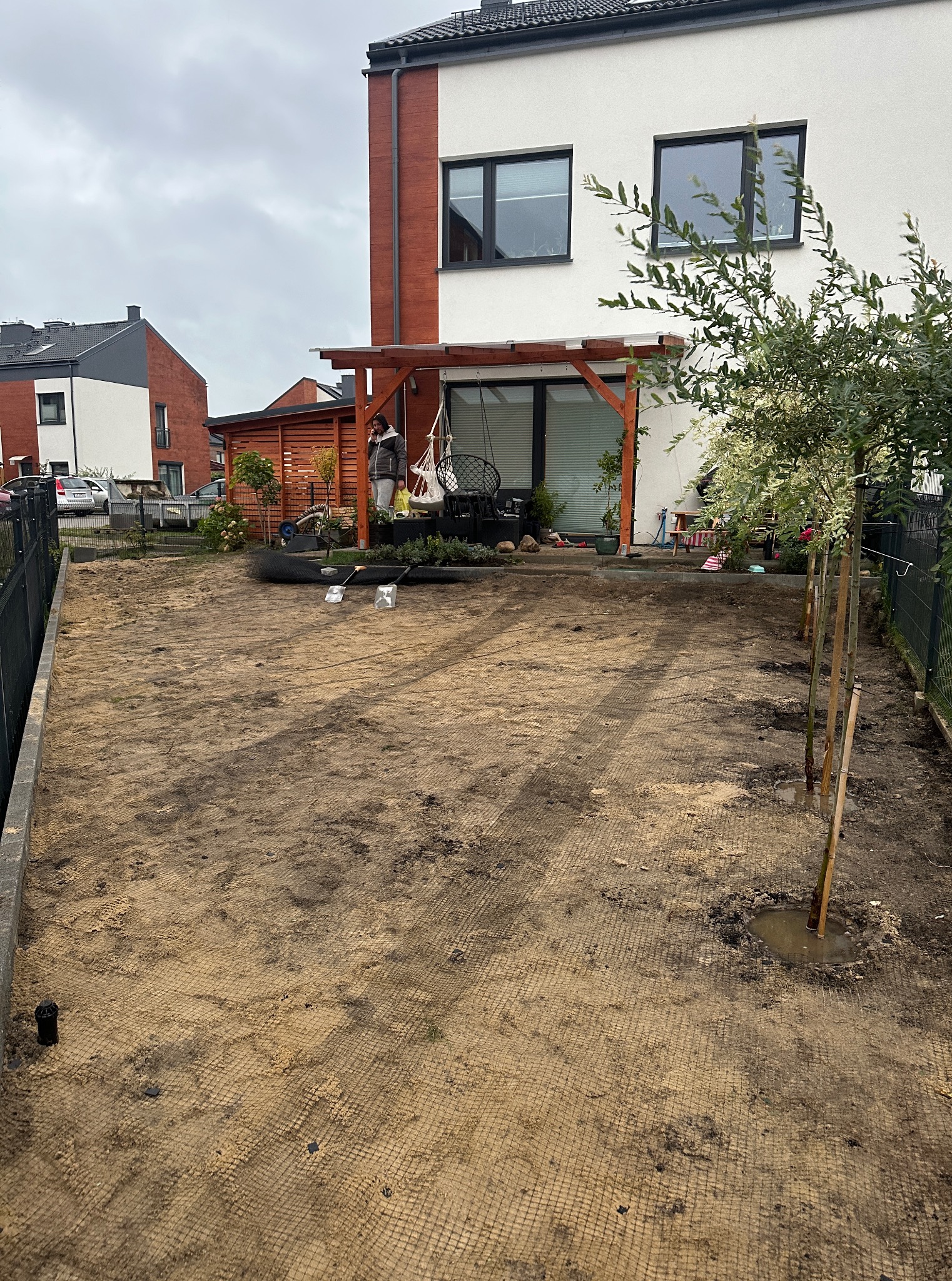 Przygotowany teren pod trawnik w Koleczkowie. Siatka wzmacniająca, młode drzewka wzdłuż ogrodzenia. W tle dom z pergolą i elementy wyposażenia ogrodu.