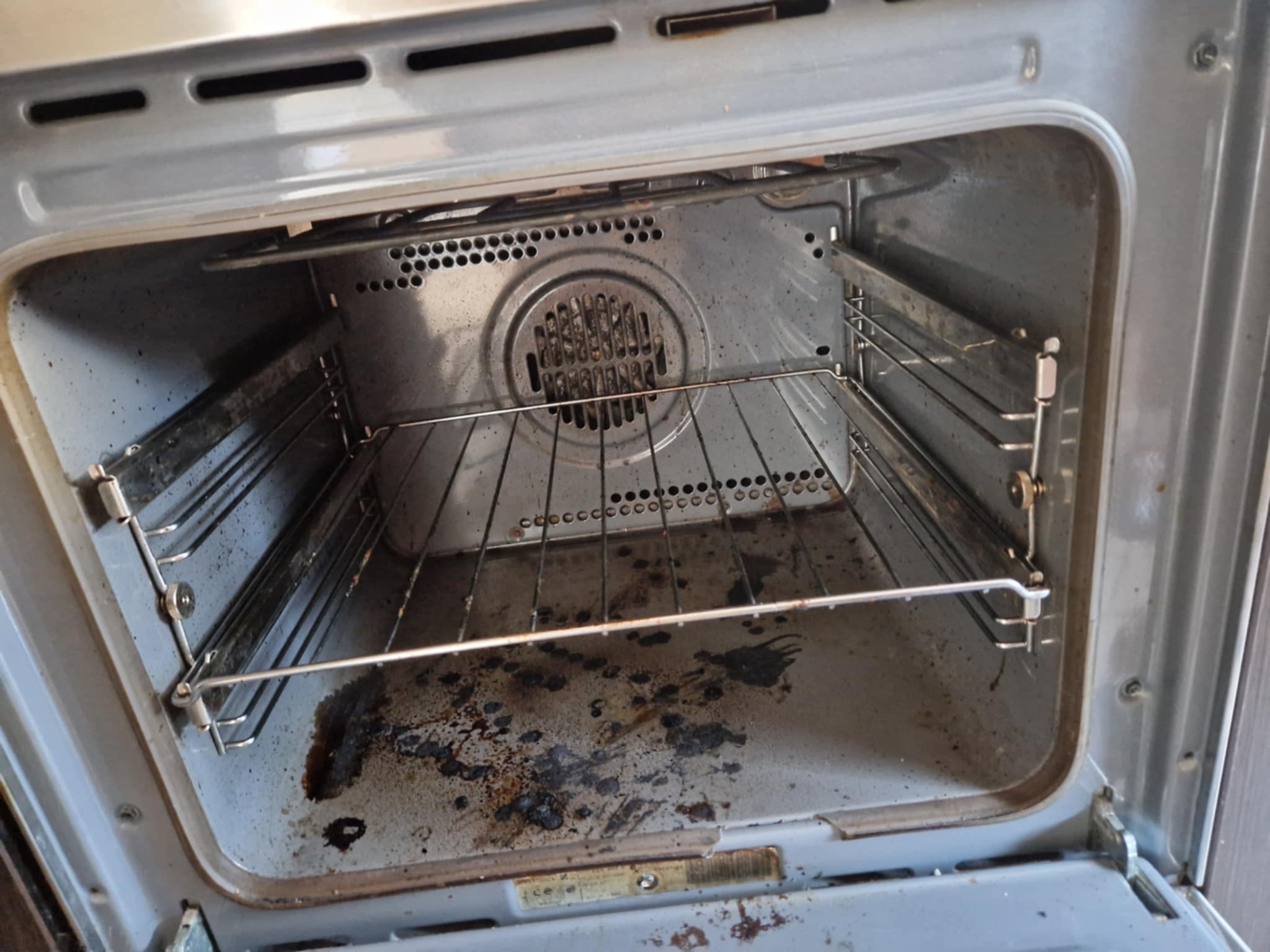 Wnętrze brudnego piekarnika z rusztem, silne zabrudzenia tłuszczem i resztkami jedzenia na dnie i ściankach. Metalowe elementy pokryte przypaleniami.