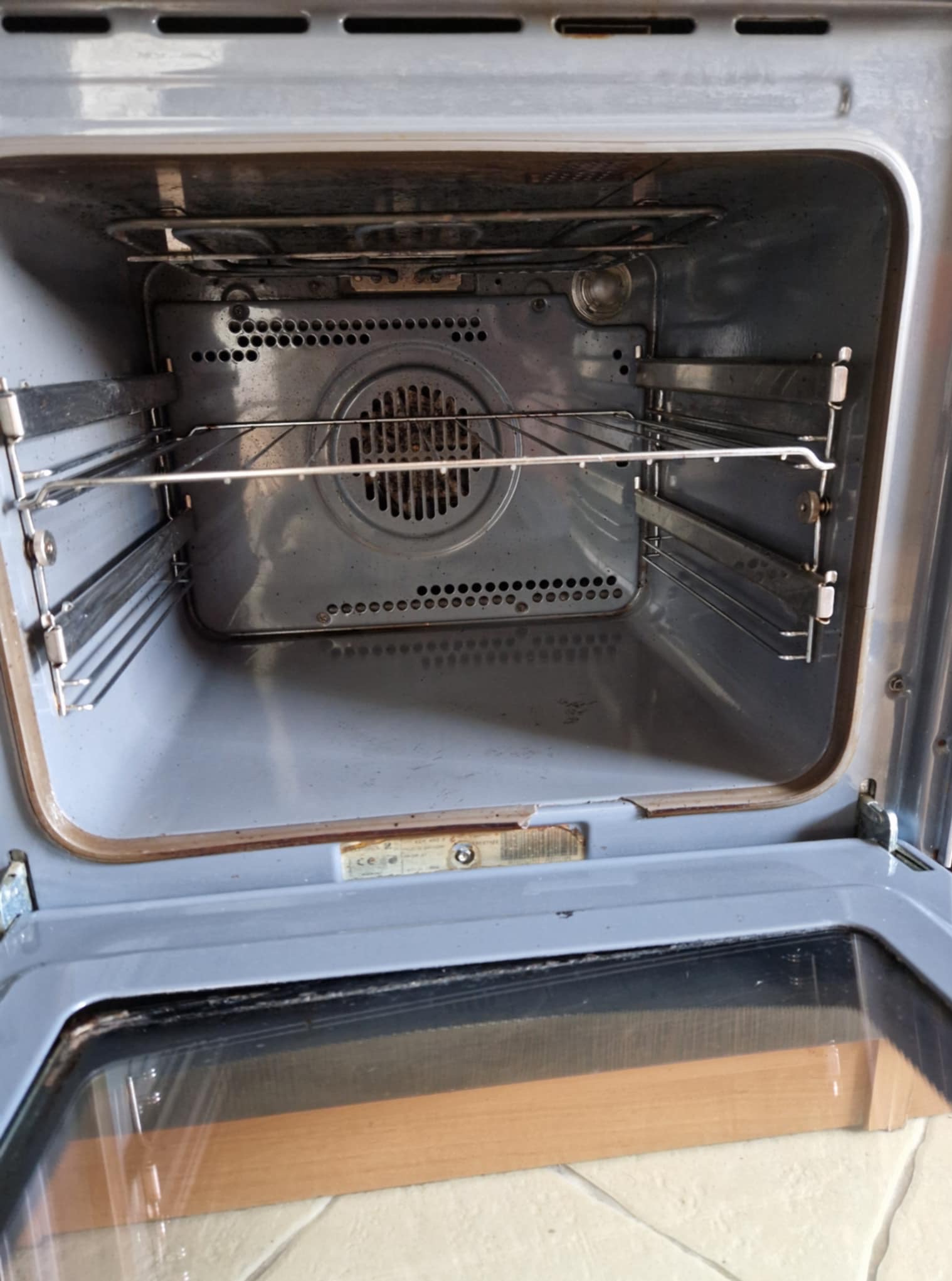 Wnętrze brudnego piekarnika przed czyszczeniem. Widoczne przypalone resztki jedzenia na ściankach i ruszcie. Otwarta klapa piekarnika z widoczną szybą.