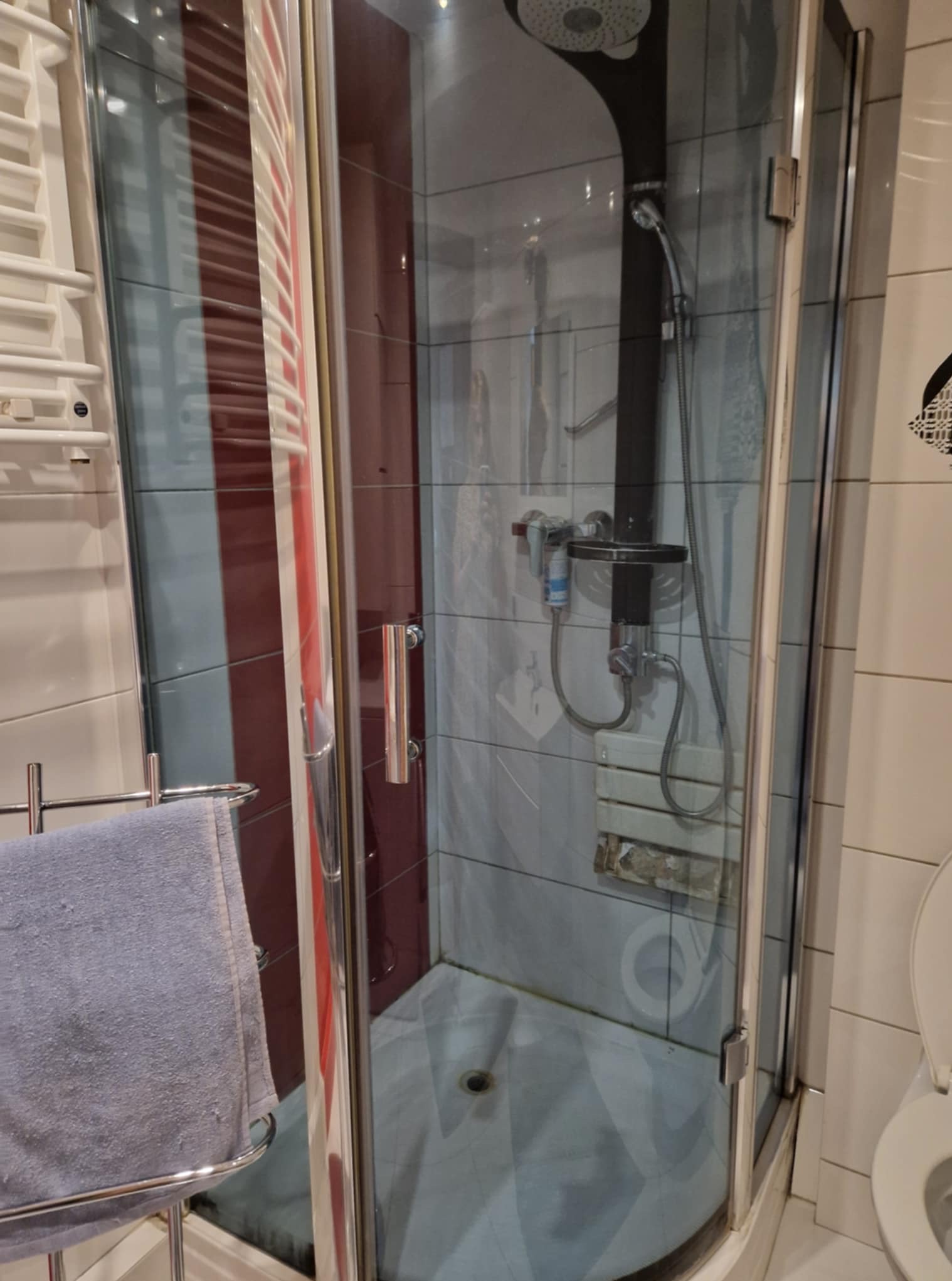 Szczegółowe ujęcie kabiny prysznicowej z widocznymi śladami użytkowania, zacieki na fugach i osad na szkle. Na wieszaku wisi szary ręcznik.