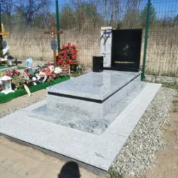 Granitowy nagrobek z czarną tablicą, ozdobiony krzyżem i kwiatami, na tle cmentarza z zieloną siatką i drzewami. Jasne, słoneczne oświetlenie.