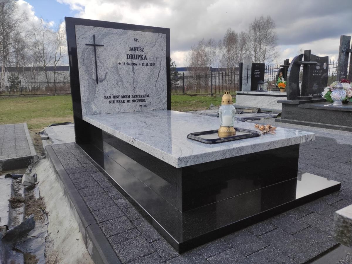 Granitowy nagrobek z czarną podstawą i jasnoszarą płytą, ozdobiony krzyżem i inskrypcją. Na płycie lampion i suszone kwiaty. Widok z boku na tle cmentarza.
