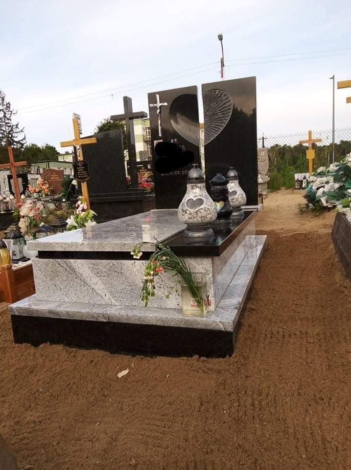 Jasnoszary nagrobek z czarnymi elementami, z dwiema pionowymi płytami z motywem serca. Na nagrobku znicze i kwiaty. W tle inne nagrobki na cmentarzu.