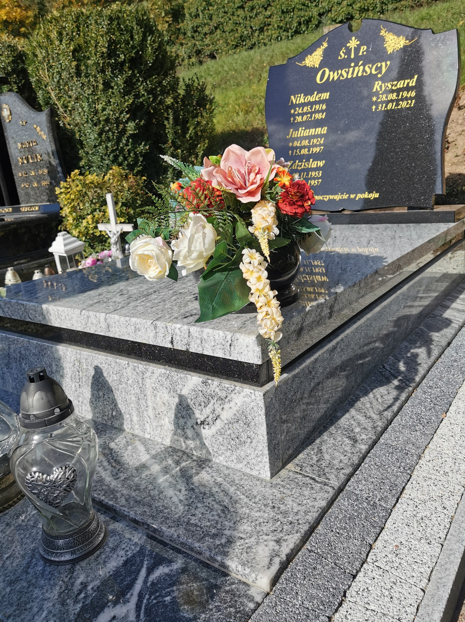 Granitowy nagrobek z czarną tablicą, zdobiony kwiatami w wazonie i lampionem ze srebrnym sercem, na tle zieleni. Widoczne detale obróbki kamienia.