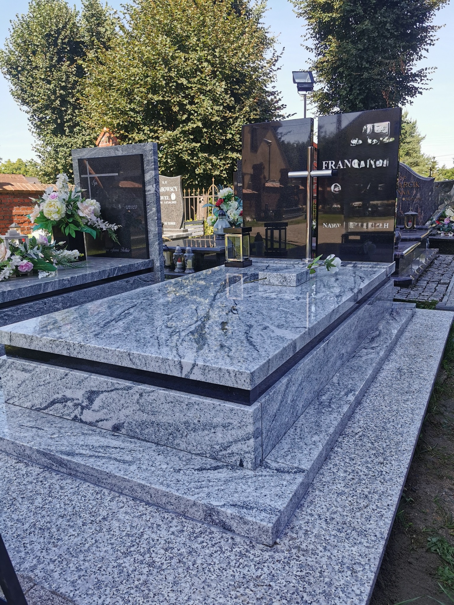 Granitowy nagrobek z jasnoszarego kamienia z ciemnymi żyłkami, z chromowanym krzyżem i kwiatami, na tle zieleni cmentarza.