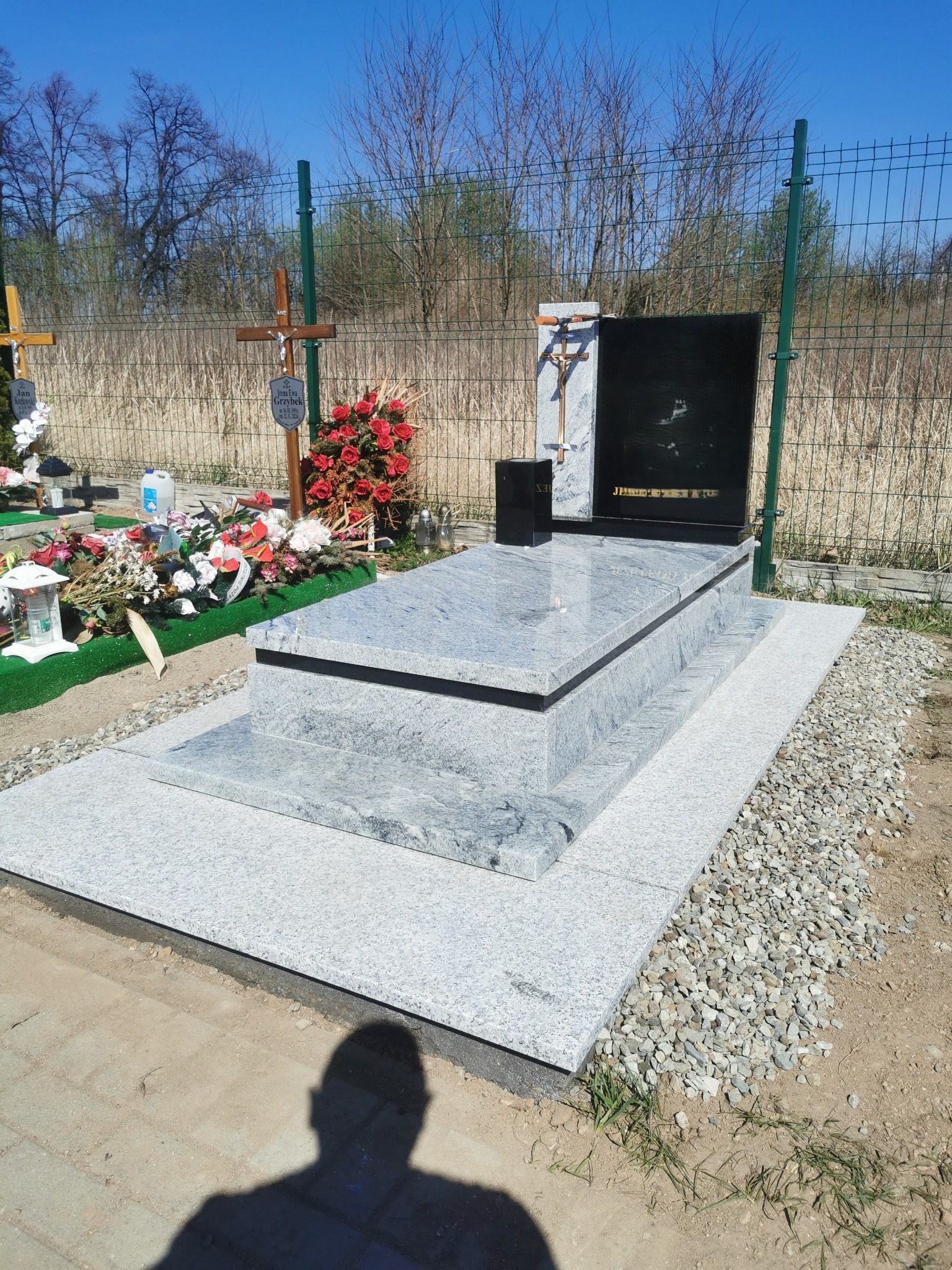 Granitowy nagrobek z czarną tablicą, ozdobiony krzyżem i kwiatami, na tle cmentarza z zieloną siatką i drzewami. Jasne, słoneczne oświetlenie.