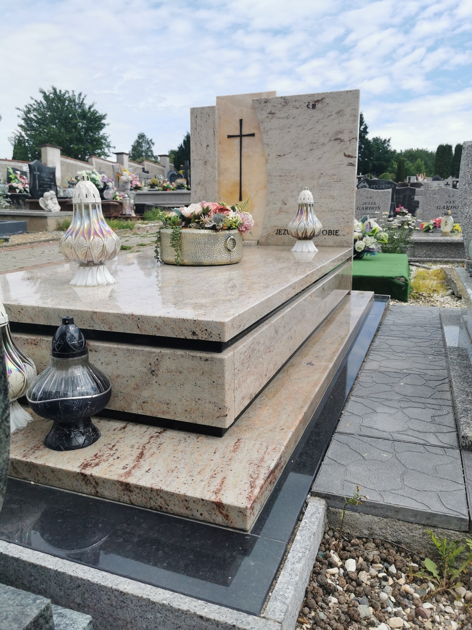 Jasny, warstwowy nagrobek z krzyżem na cmentarzu, z kwiatami i lampionami. Nagrobek wykonany z kamienia o delikatnym wzorze. Tło cmentarza z innymi nagrobkami.