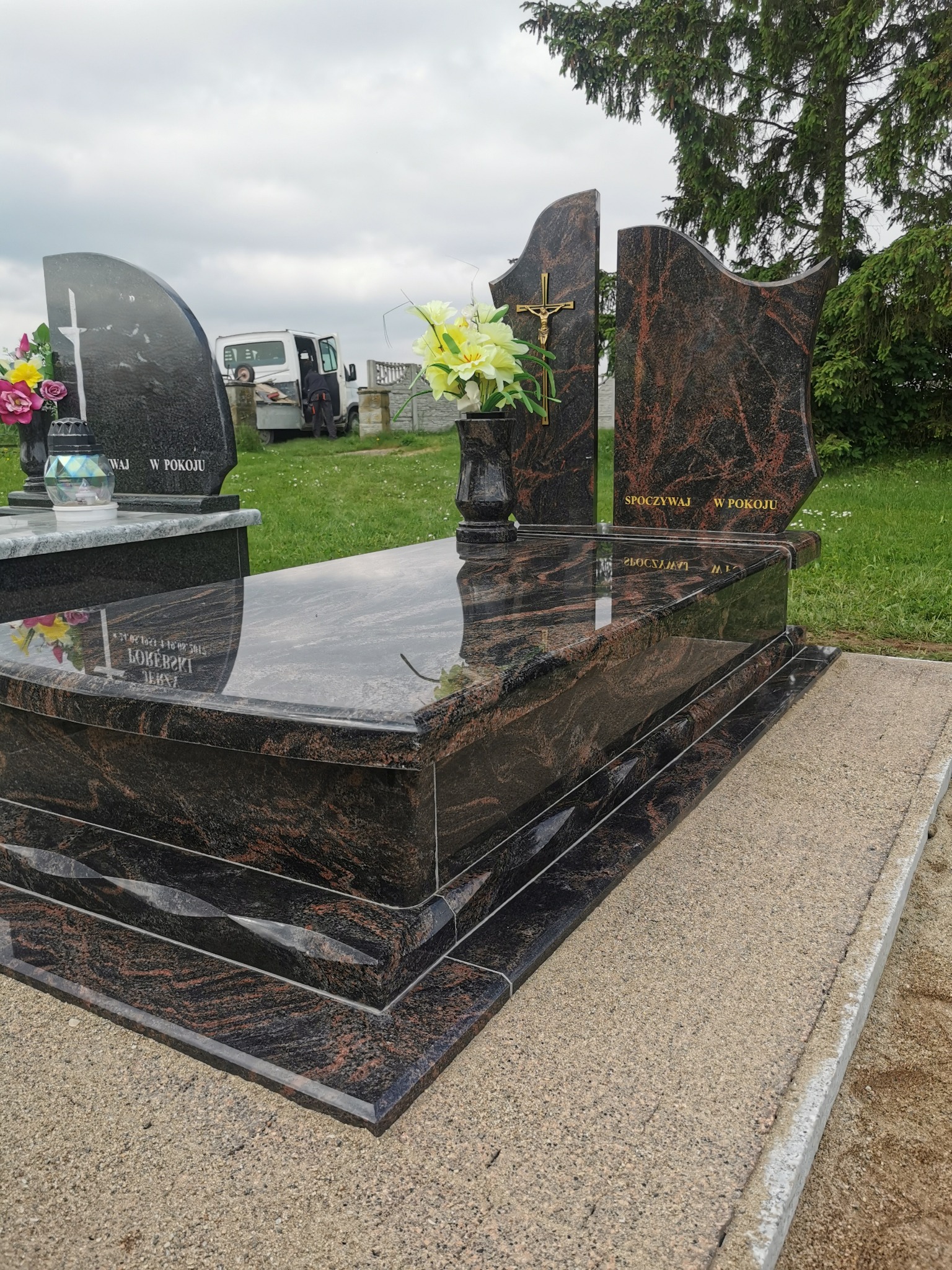 Elegancki, polerowany nagrobek z ciemnego granitu z brązowymi żyłkami, z inskrypcją i złotym krzyżem, na tle zieleni i pochmurnego nieba.