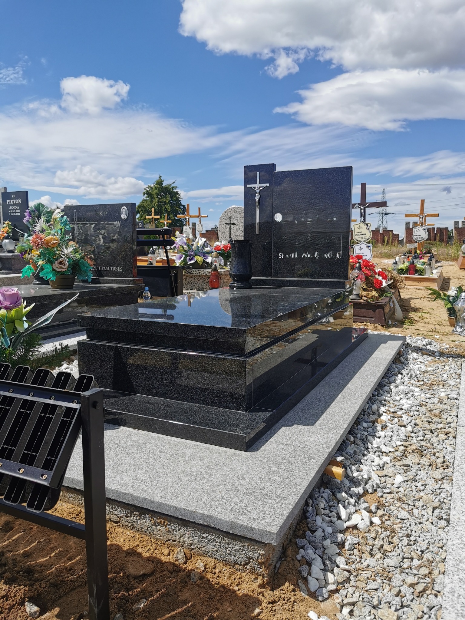 Czarny, błyszczący nagrobek z krzyżem na cmentarzu w słoneczny dzień. Widoczne inne nagrobki i błękitne niebo z chmurami w tle. Nagrobek otoczony kamieniami.