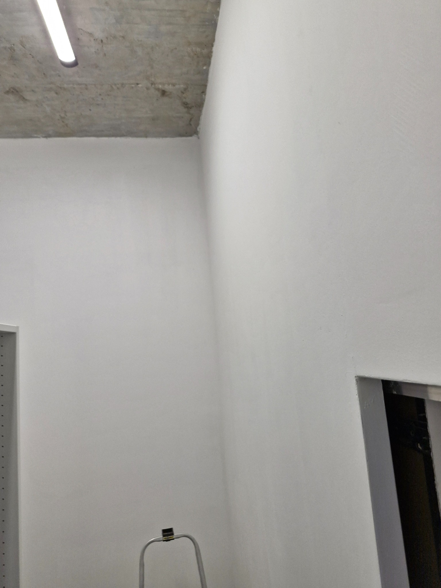 Pomieszczenie po malowaniu na biało, widoczny fragment surowego sufitu, ściany, fragment regału i drabiny malarskiej.
