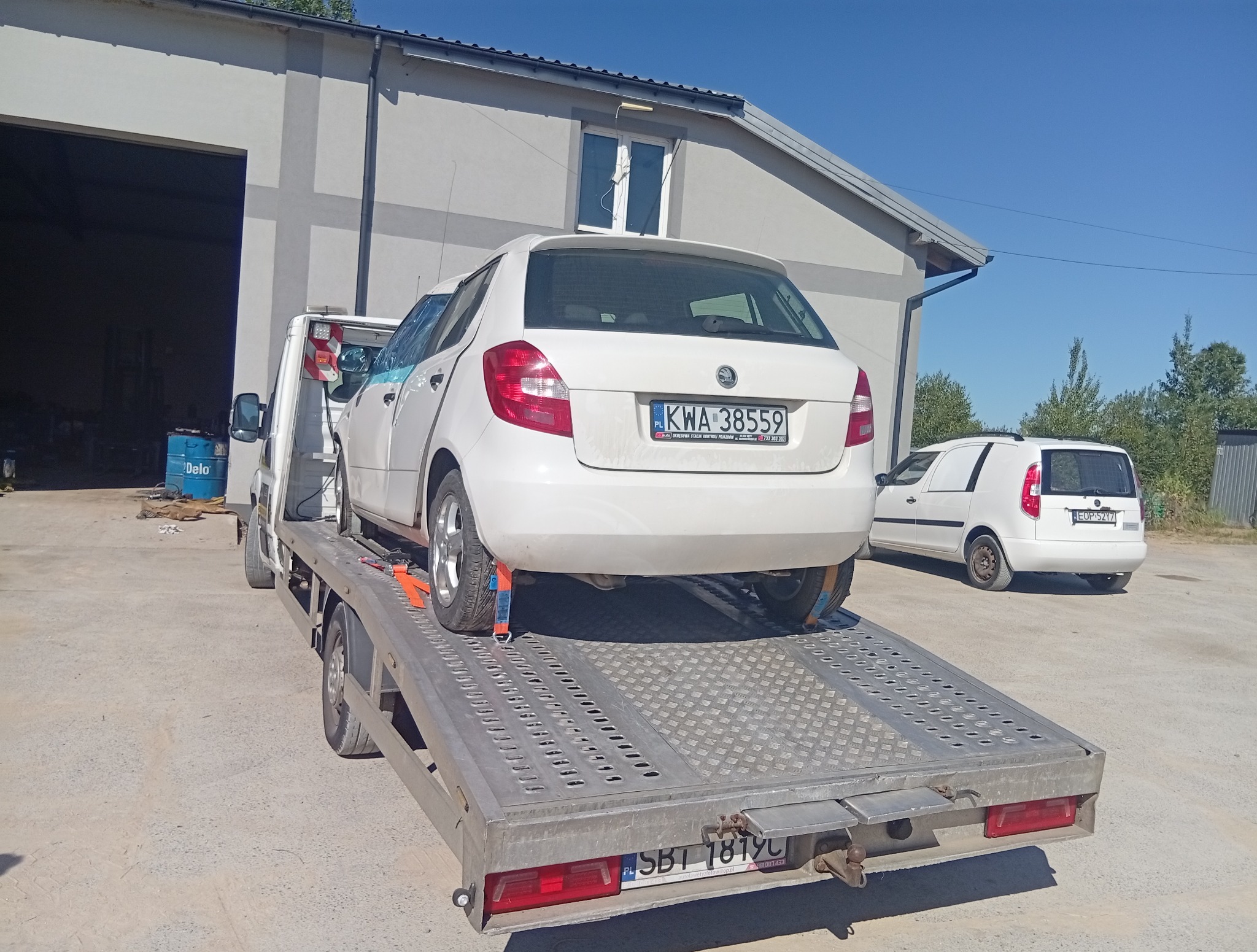 Biała Skoda Fabia zabezpieczona pasami na platformie lawety. W tle budynek z otwartą bramą i biały bus. Słoneczny dzień.