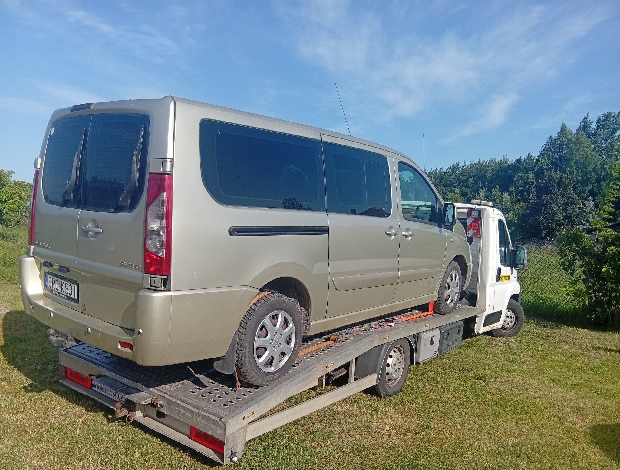 Bus Peugeot Expert na lawecie, widok z tyłu, tablica rejestracyjna widoczna. Laweta na trawie, w tle zieleń i błękitne niebo. Transport pojazdów.