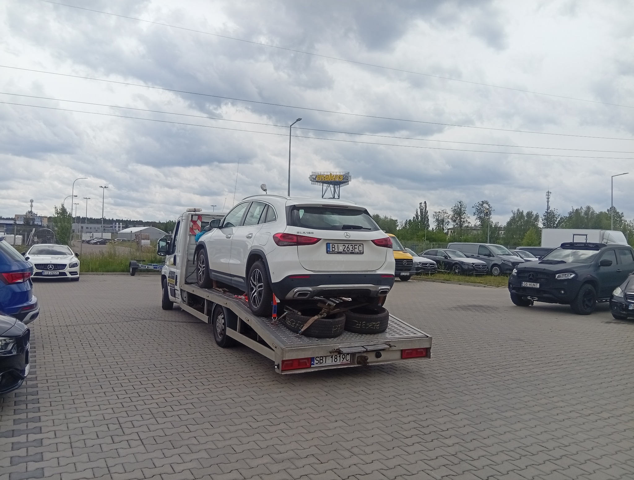 Biały Mercedes GLA na lawecie, widok z tyłu, na parkingu z innymi autami. Tablice rejestracyjne widoczne. Dodatkowe opony na platformie. Pochmurne niebo w tle.