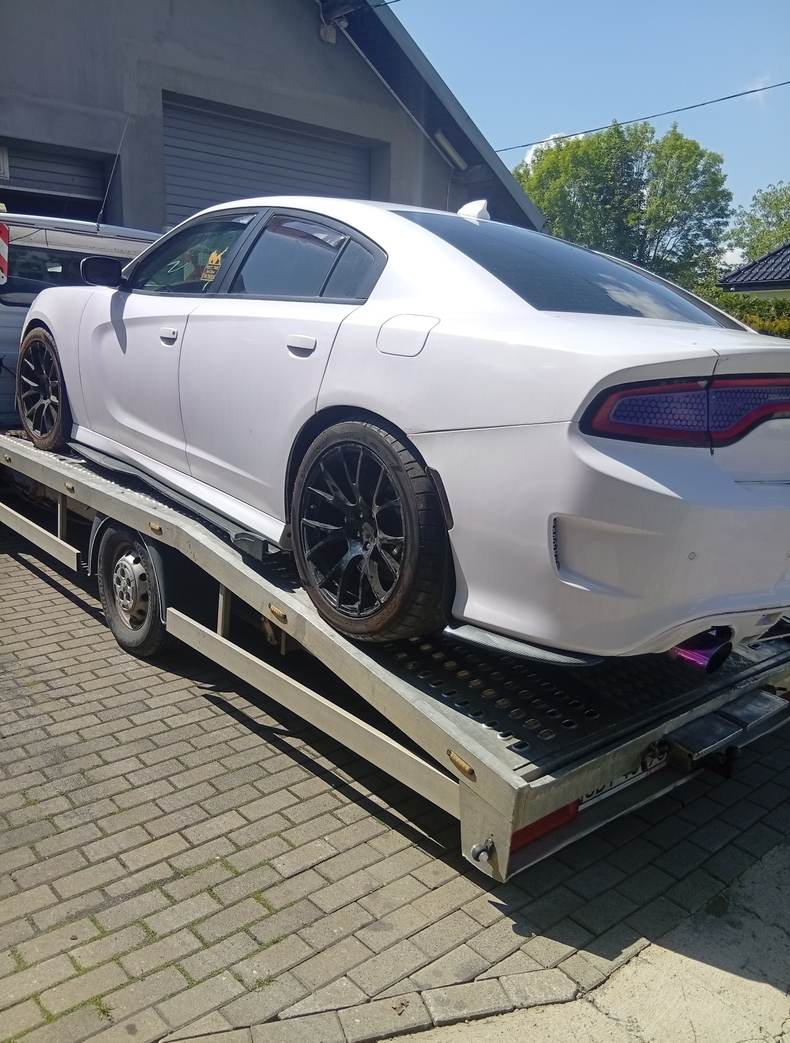 Biały Dodge Charger z czarnymi felgami na srebrnej lawecie, widok z tyłu. Fioletowy wydech. Naklejka na szybie. Kostka brukowa w tle.