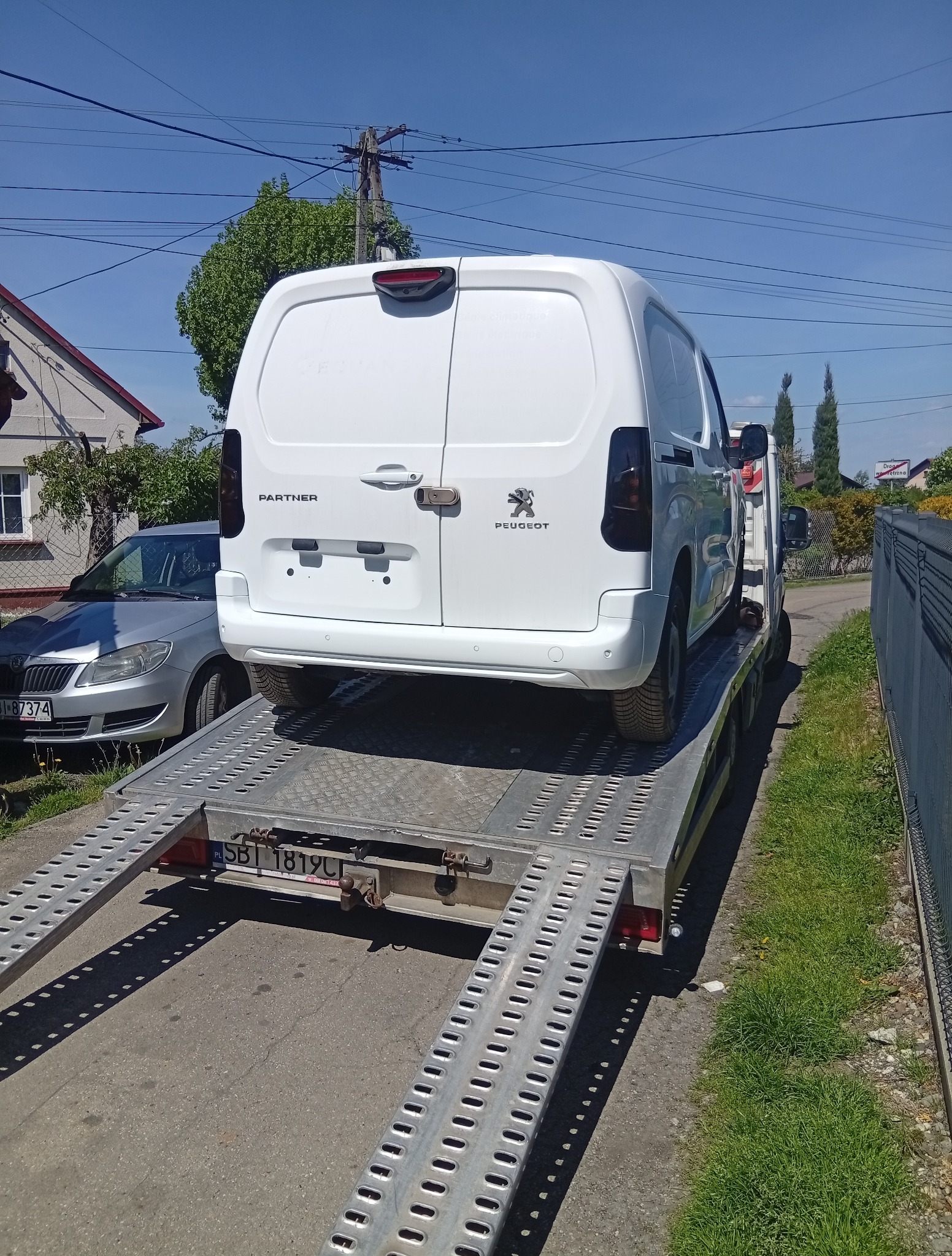 Biały Peugeot Partner na lawecie, widok z tyłu na ulicy. Obok zaparkowany srebrny samochód osobowy, w tle domy i zieleń. Laweta z polską tablicą rejestracyjną.