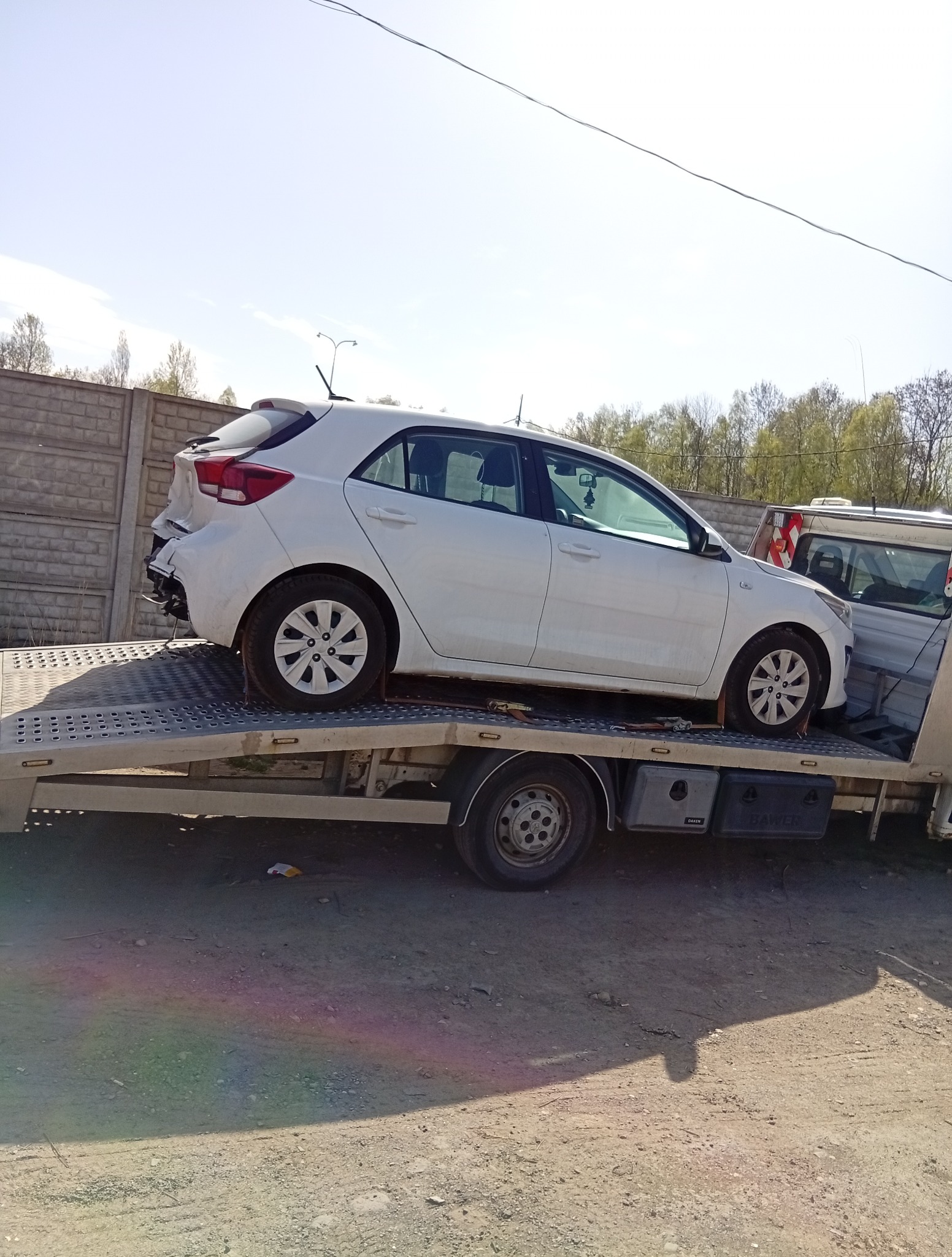 Biały uszkodzony samochód na lawecie. Widoczne uszkodzenia tylnej części pojazdu. Laweta zaparkowana na żwirowym podłożu, w tle betonowy płot i drzewa.