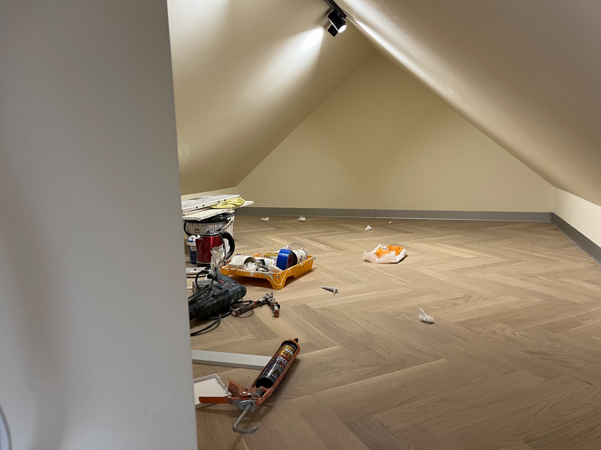 Poddasze po malowaniu ścian, z narzędziami malarskimi na parkiecie w jodełkę. Widoczne puszki z farbą, wałek i pistolet do silikonu.