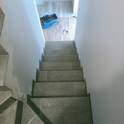 Usługi Ogólnobudowlane Dawid Pranga - Wykończone płytkami schody wewnątrz budynku, widok z góry. Świeżo pomalowane ściany. Na podłodze wiadro z narzędziami do fugowania.