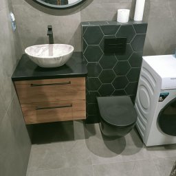 Usługi Ogólnobudowlane Dawid Pranga - Nowoczesna łazienka z umywalką marmurkową na szafce z drewnianymi szufladami, toaleta w zabudowie z heksagonalnymi płytkami i pralka. Szare płytki na ścianach i podłodze.