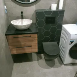 Nowoczesna łazienka z umywalką marmurkową na szafce z drewnianymi szufladami, toaleta w zabudowie z heksagonalnymi płytkami i pralka. Szare płytki na ścianach i podłodze.