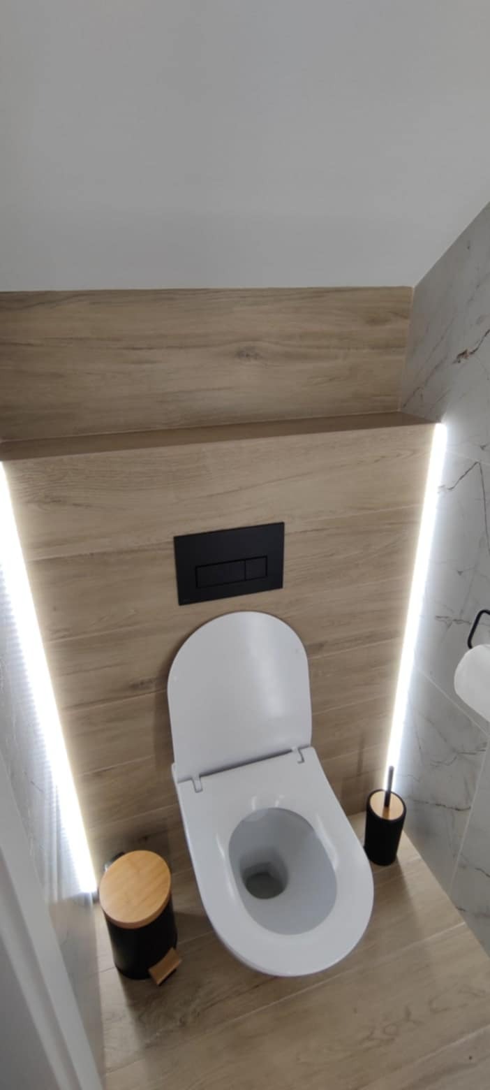 Nowoczesna toaleta podwieszana z drewnopodobną okładziną, czarnym przyciskiem spłukującym i subtelnym oświetleniem LED w wykończonej łazience.