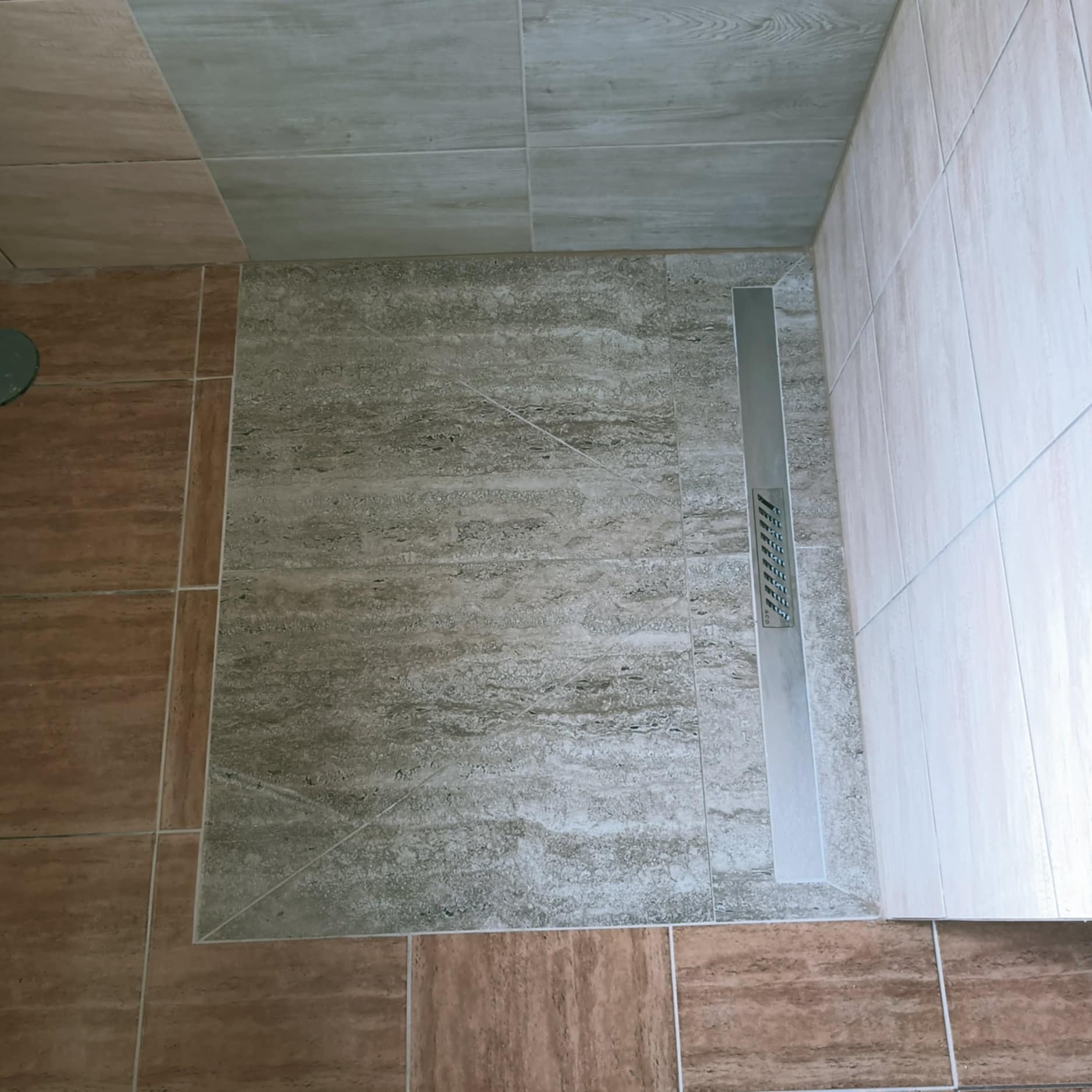 Wykończona strefa prysznicowa z odpływem liniowym i płytkami imitującymi kamień w odcieniach szarości i beżu. Widok z góry na fragment łazienki po remoncie.