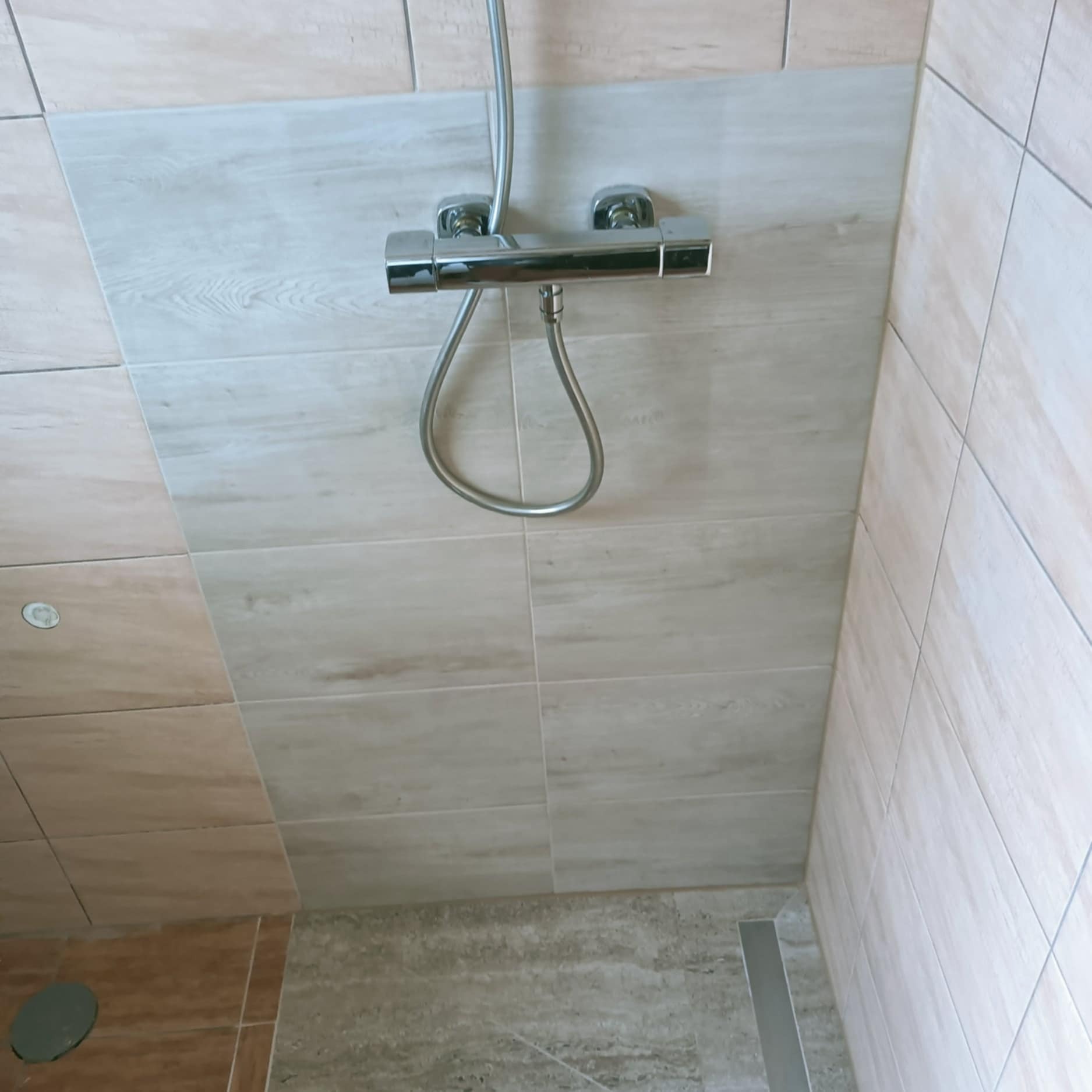 Wykończona kabina prysznicowa z chromowaną armaturą i beżowymi płytkami imitującymi drewno. Widoczny odpływ liniowy. Minimalistyczny design.