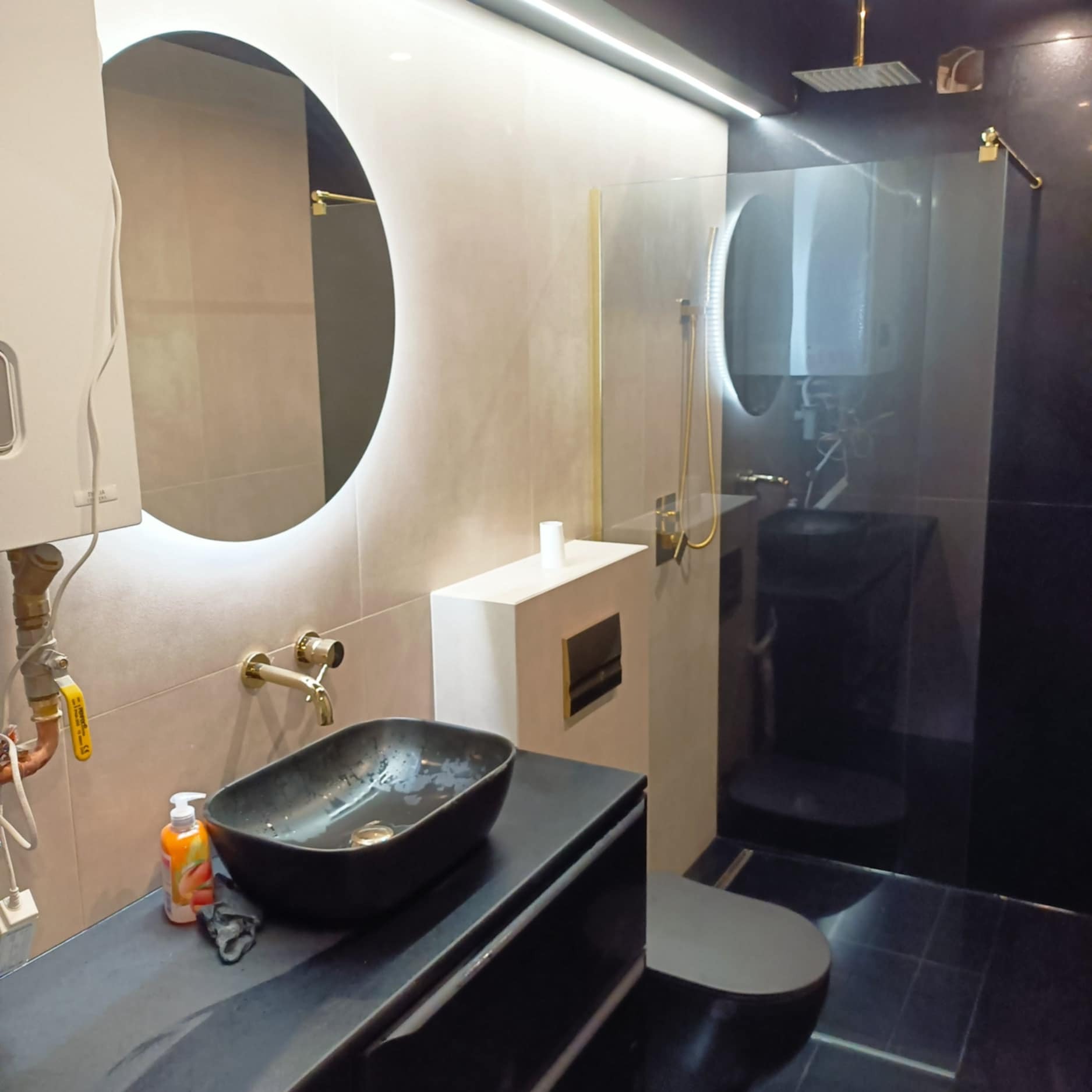 Nowoczesna łazienka z czarną umywalką, okrągłym lustrem z podświetleniem LED i kabiną prysznicową typu walk-in z złotymi akcentami.