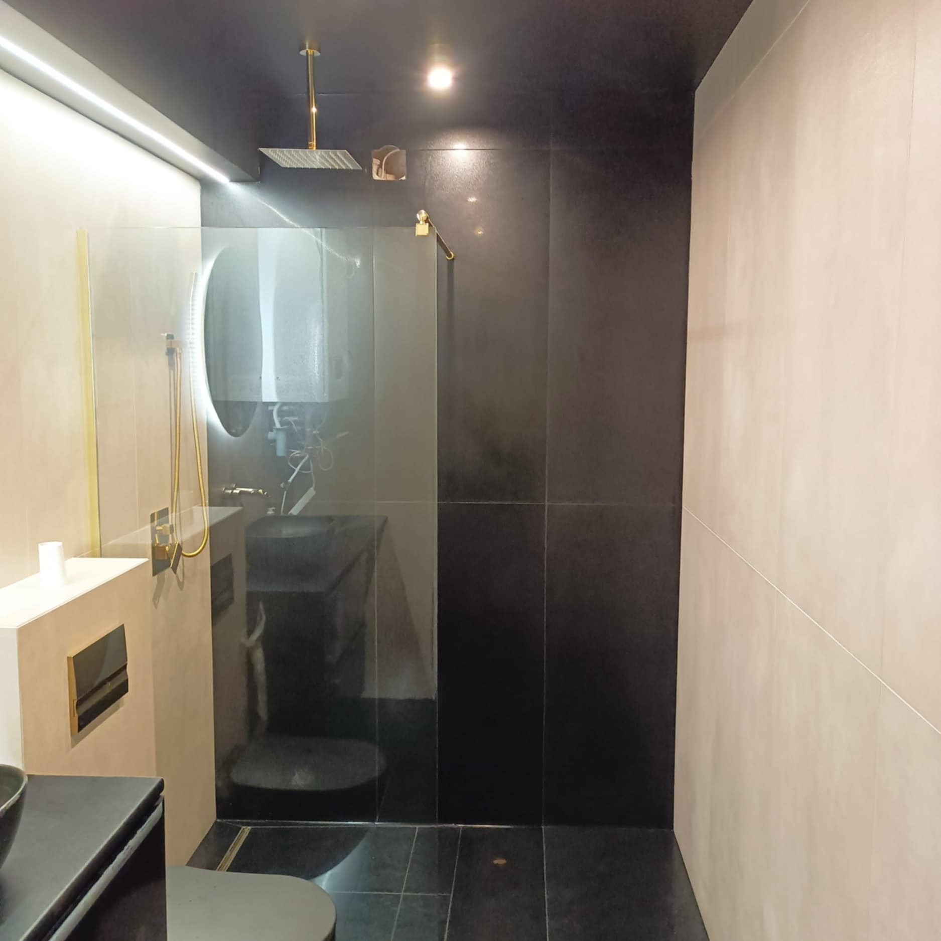 Nowoczesna łazienka z czarnymi i beżowymi płytkami, złotą armaturą i oświetleniem LED. Kabina prysznicowa ze szklaną ścianką i okrągłym lustrem z podświetleniem.