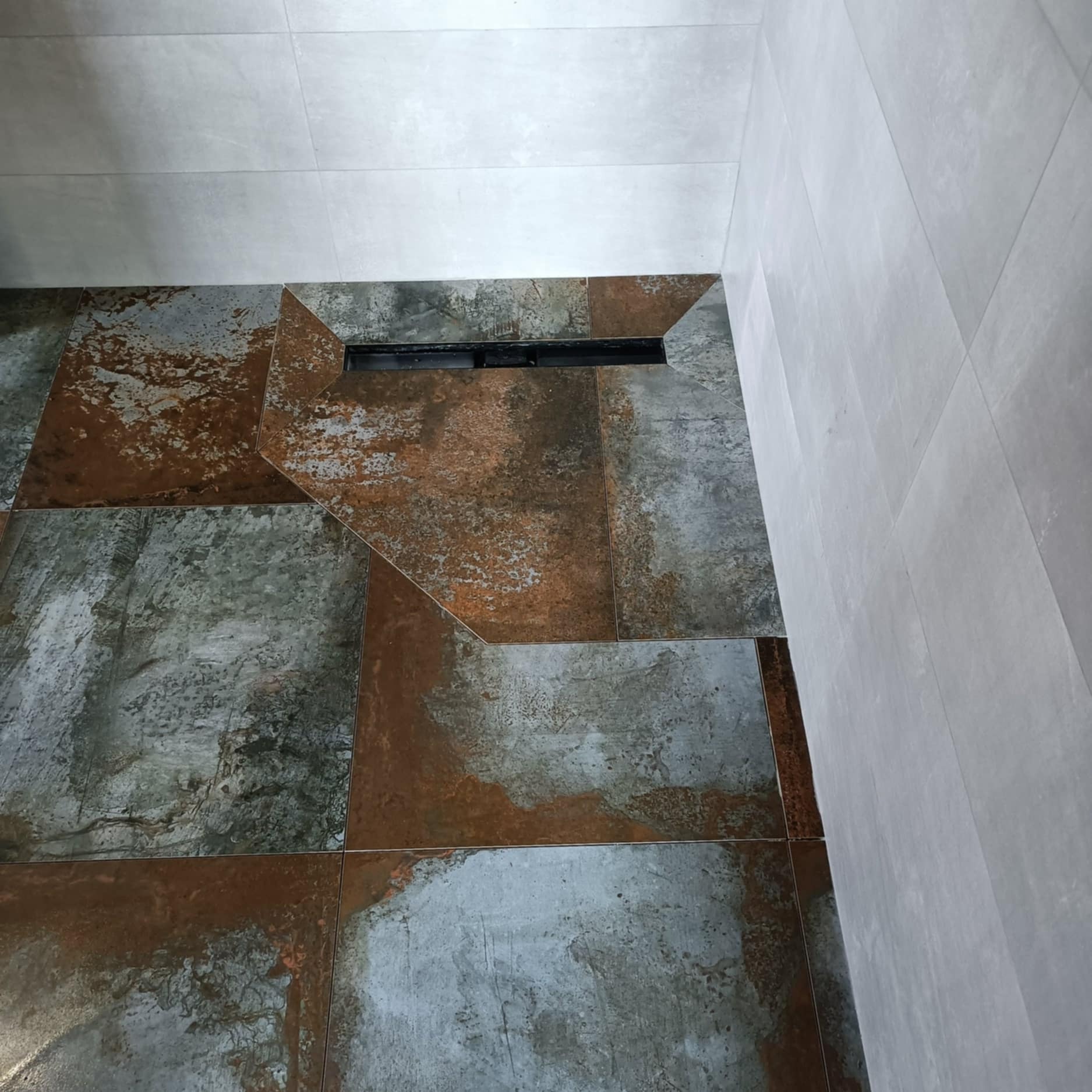 Nowoczesna łazienka z rustykalnymi płytkami podłogowymi w odcieniach szarości i rdzy, oraz minimalistycznym odpływem liniowym. Ściany wyłożone jasnymi, gładkimi płytkami.