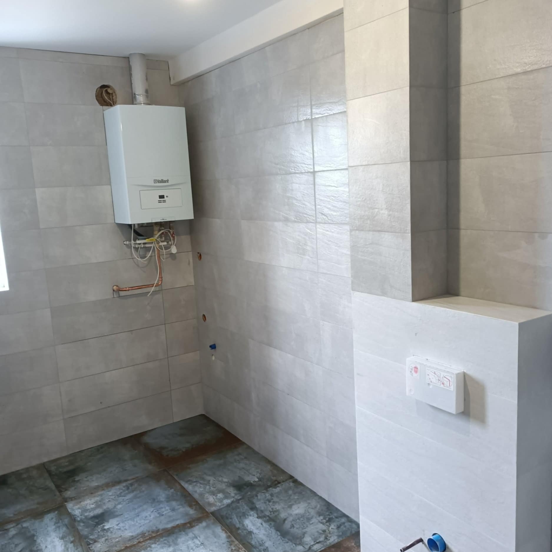 Wykończona łazienka z szarymi płytkami, wiszącym bojlerem i zabudowaną spłuczką. Widoczne przyłącza wodne i kanalizacyjne. Minimalistyczny design.